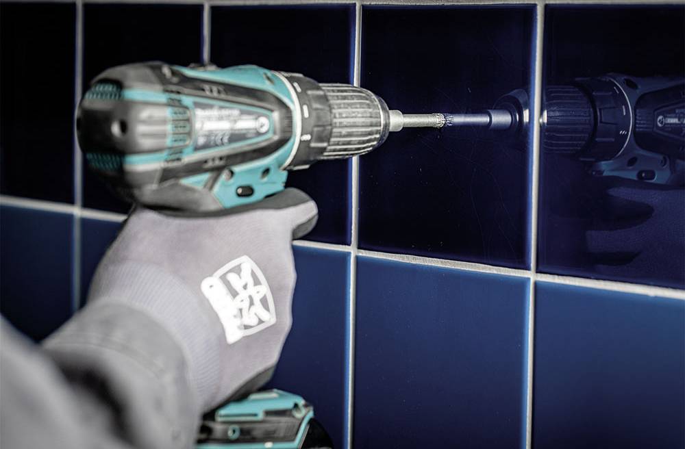 Osoba wiertli akumulatorowym wkrętakiem otwór w ścianie pokrytej niebieskimi kafelkami. Osoba ma na ręce rękawicę.