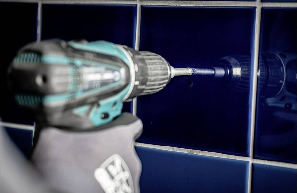 Osoba wierca otwór akumulatorowym wkrętakiem w niebieskim kafelku. Akcent położony jest na narzędziu i wykonywanej czynności.