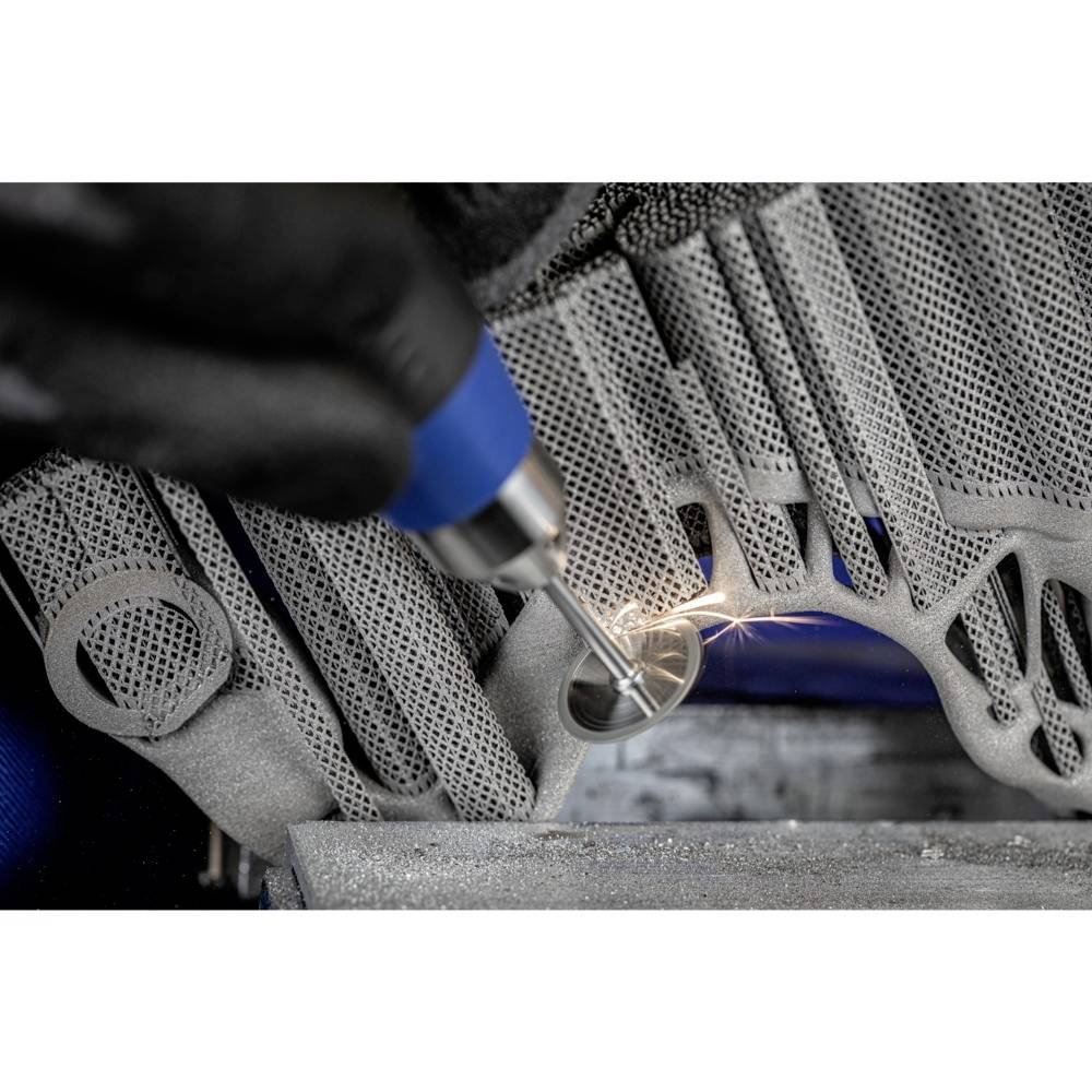 Osoba obrabia szary element metalowy niebieskim narzędziem ręcznym, przy czym wytwarzają się iskry. Nacisk kładziony jest na precyzyjną pracę warsztatową.