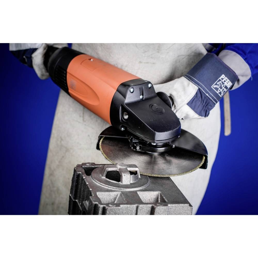 Osoba ubrana w strój roboczy obsługuje szlifierkę kątową przy metalowym bloku na niebieskim tle.