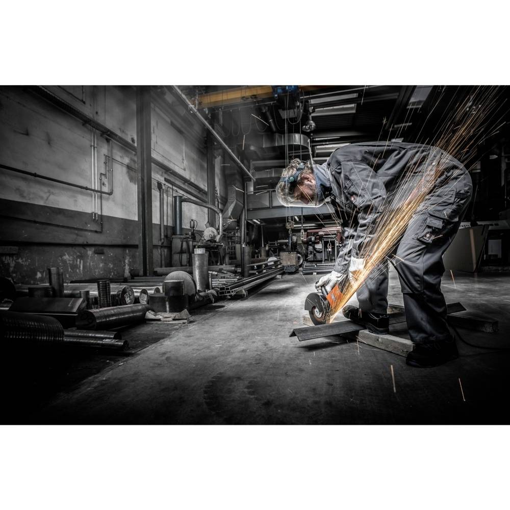 Osoba w odzieży ochronnej szlifuje metal w warsztacie, iskry lecą. Przemysłowe otoczenie z maszynami i rurami.