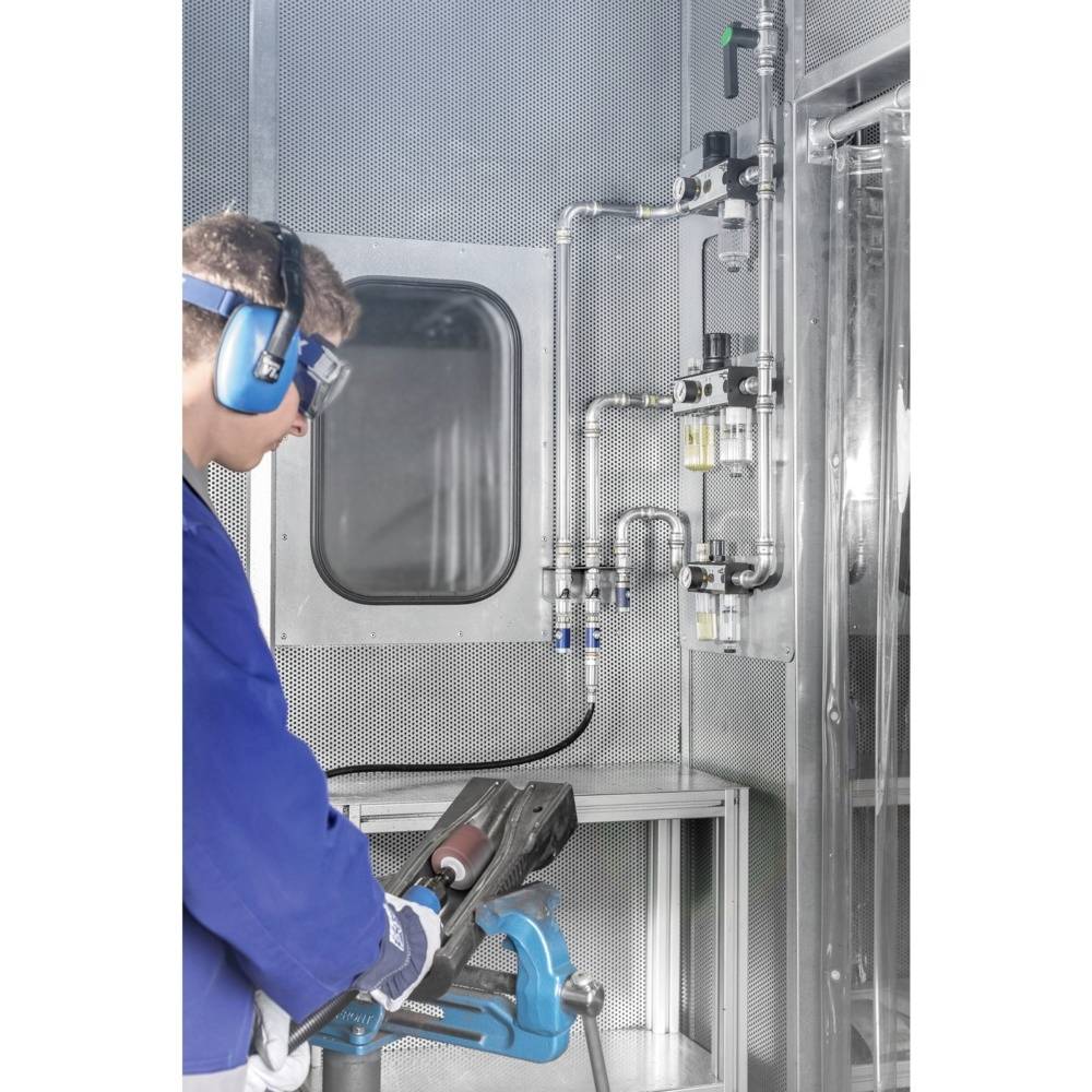 Osoba w stroju ochronnym pracuje w pomieszczeniu technicznym z zaworami i rurami. Nosi ochronniki słuchu i okulary ochronne.