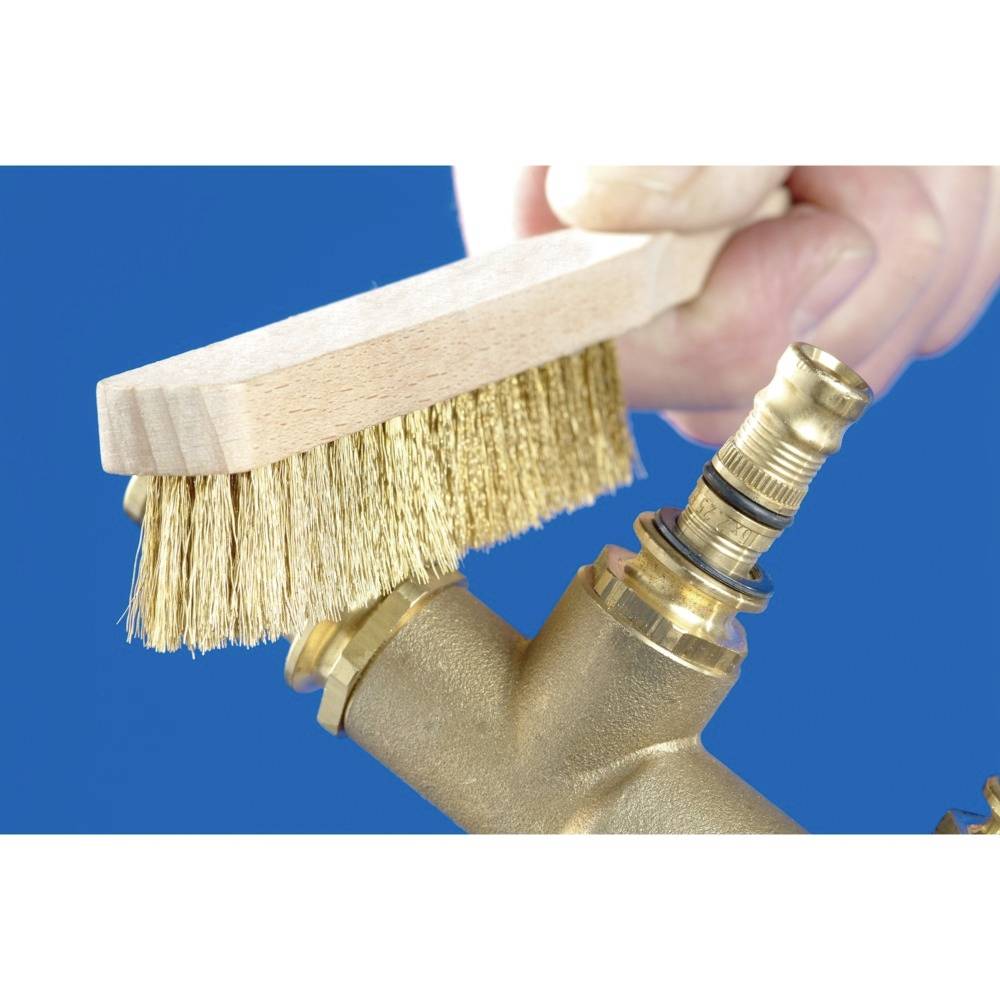 Dłoń trzyma szczotkę drucianą i czyści mosiężną rurę na niebieskim tle.