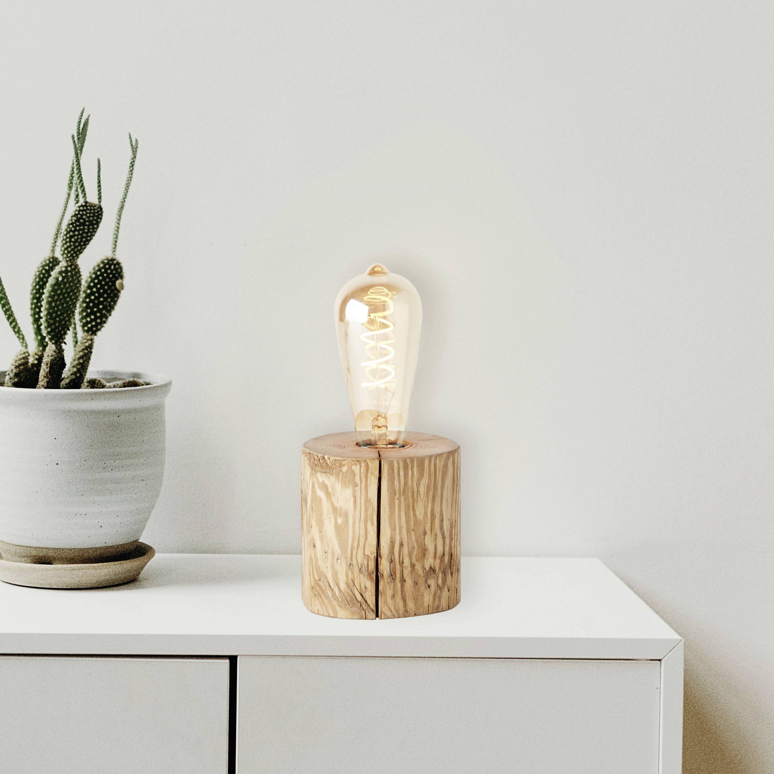 Żarówka na drewnianym stojaku świeci obok kaktusa w glinianej doniczce. Oba stoją na białej szafce.