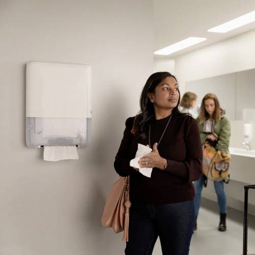 Osoba osusza ręce papierowym ręcznikiem w publicznej toalecie. W tle widać dwie kolejne osoby.