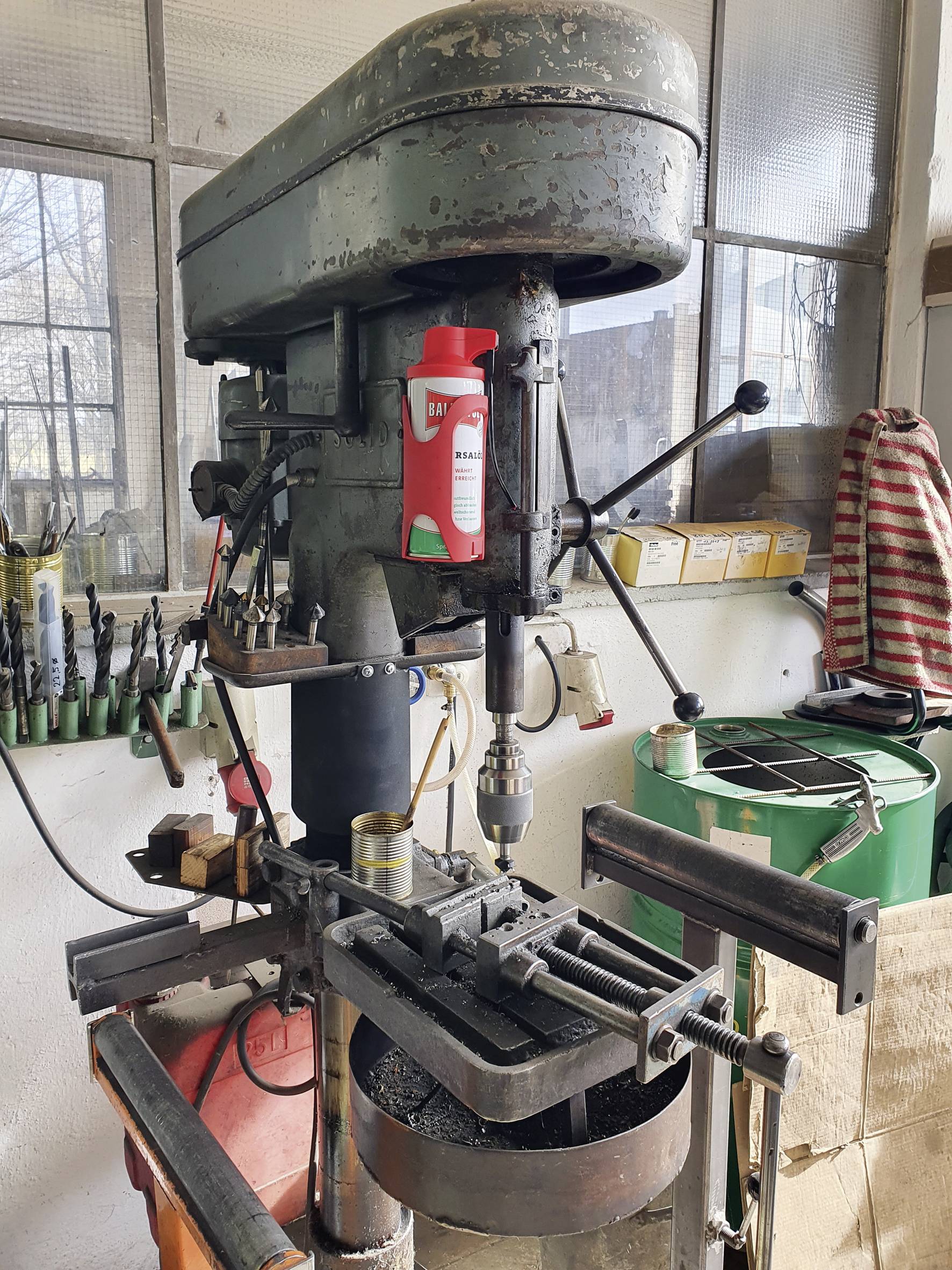 Stara wiertarka stojąca w warsztacie, otoczona narzędziami. Czerwona butelka z olejem stoi na jej wierzchu. W tle drewno i skrzynie.