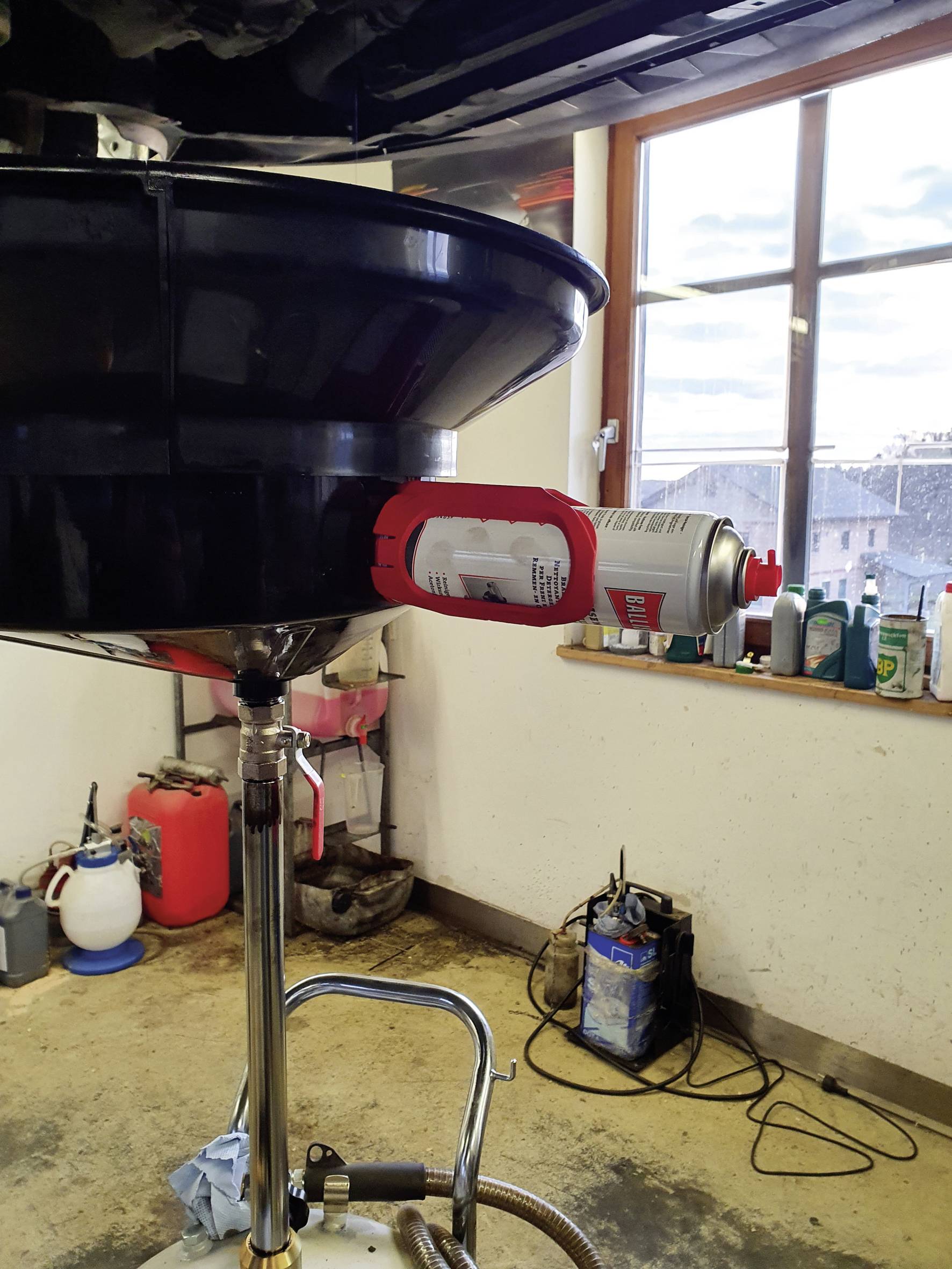 Warsztat przedstawia urządzenie do wymiany oleju z dozownikiem sprayu przymocowanym z boku. W tle widoczne są okna i narzędzia.