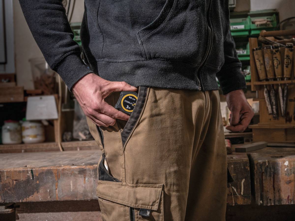 Osoba w warsztacie wkłada miarkę do kieszeni spodni. W tle widoczne są narzędzia i stół roboczy.