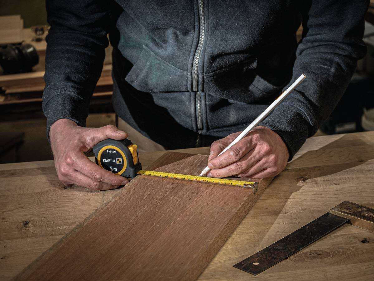 Osoba mierzy drewnianą deskę za pomocą miarki i zaznacza ją ołówkiem. Narzędzia leżą na stole.