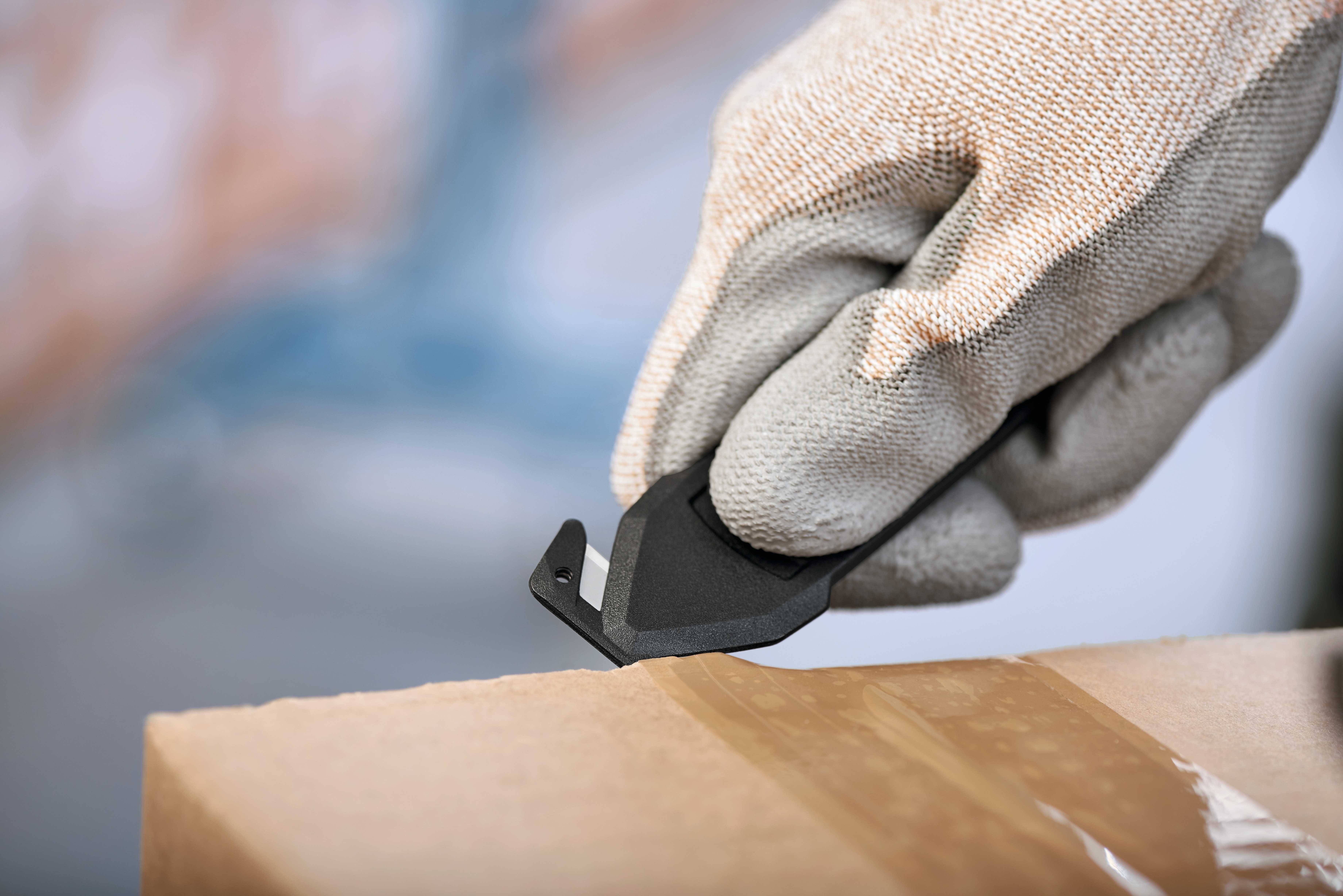 Dłoń w rękawicy kroi taśmę klejącą na kartonie przy pomocy noża bezpieczeństwa.