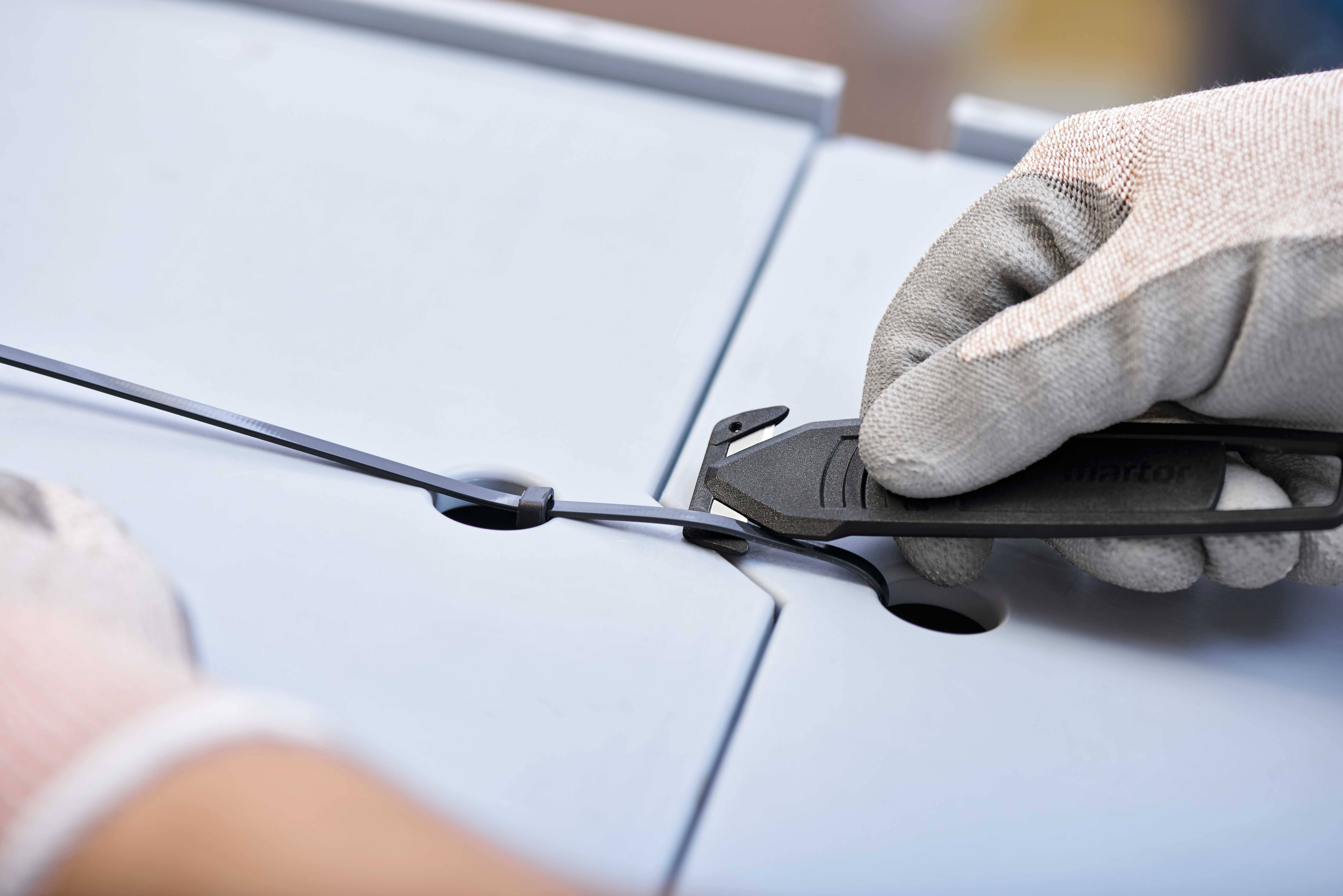 Osoba w rękawiczkach przecina nożem opaskę zaciskową łączącą dwie szare płyty.