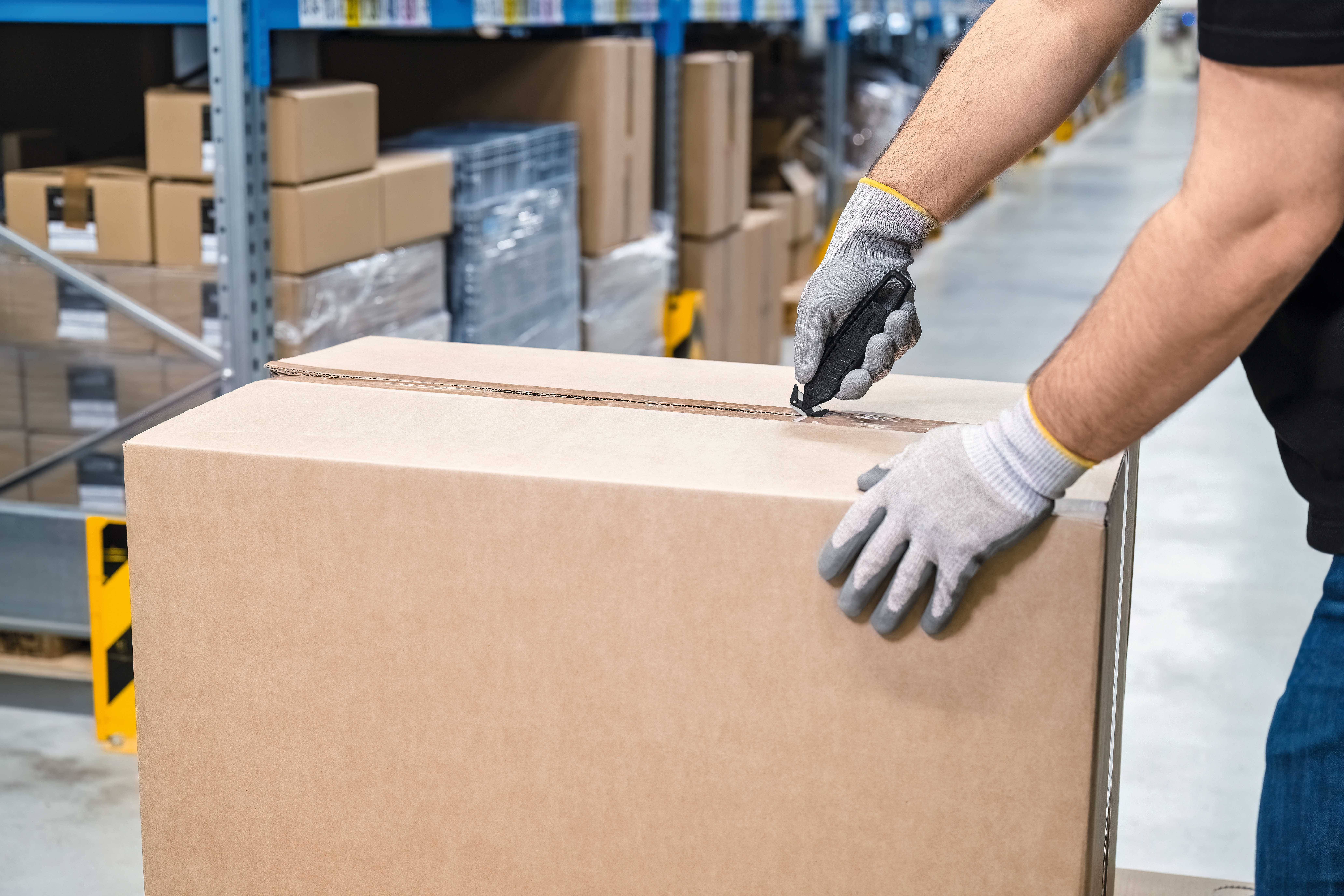 Osoba otwiera duży karton za pomocą noża introligatorskiego w magazynie, otoczona regałami pełnymi kolejnych paczek i kartonów.