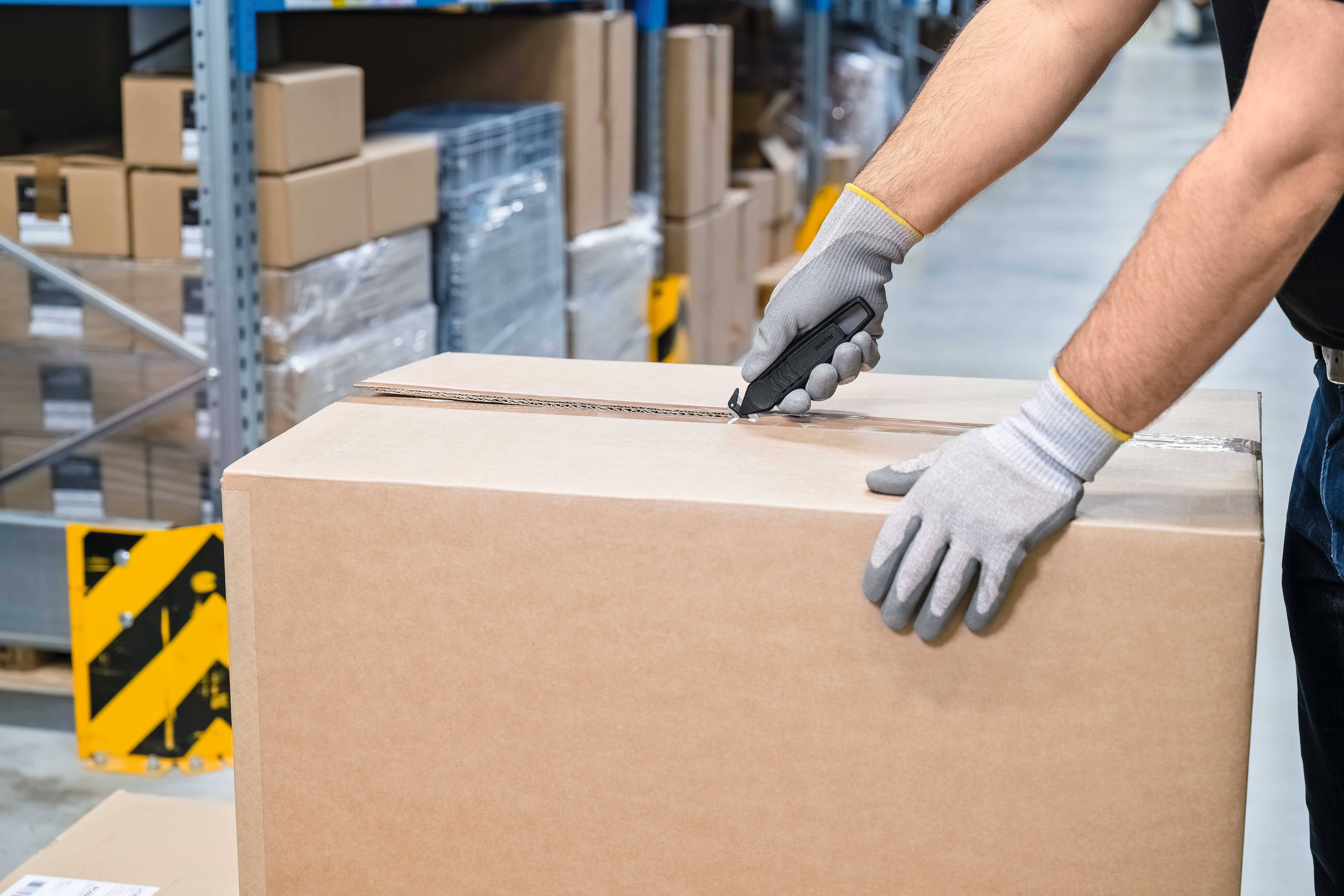 Osoba otwiera karton nożem do tapet w magazynie. Ręce są w rękawicach. W tle widać regały z towarami.