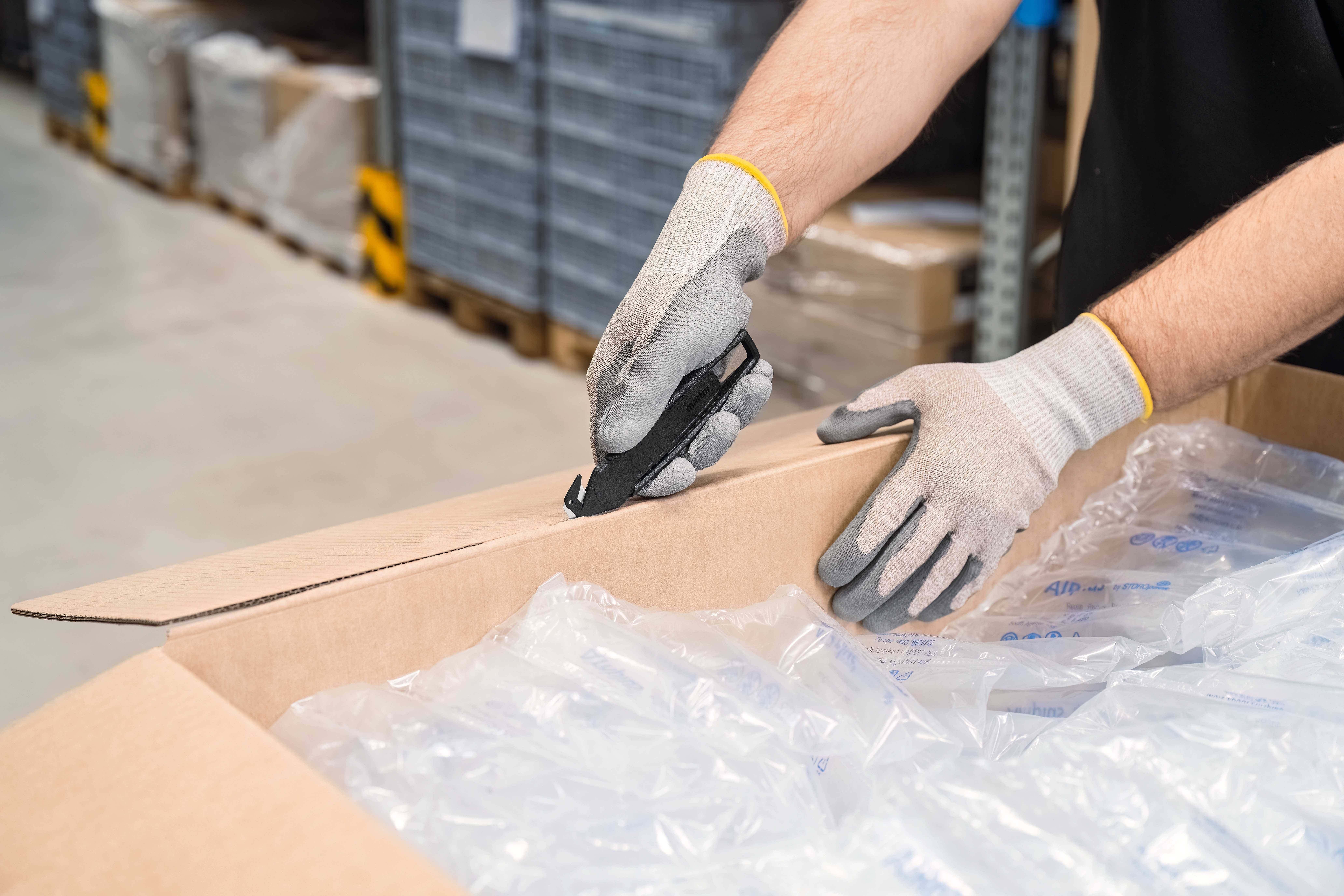 Osoba w rękawiczkach otwiera nożem karton w magazynie, wypełniony folią bąbelkową.