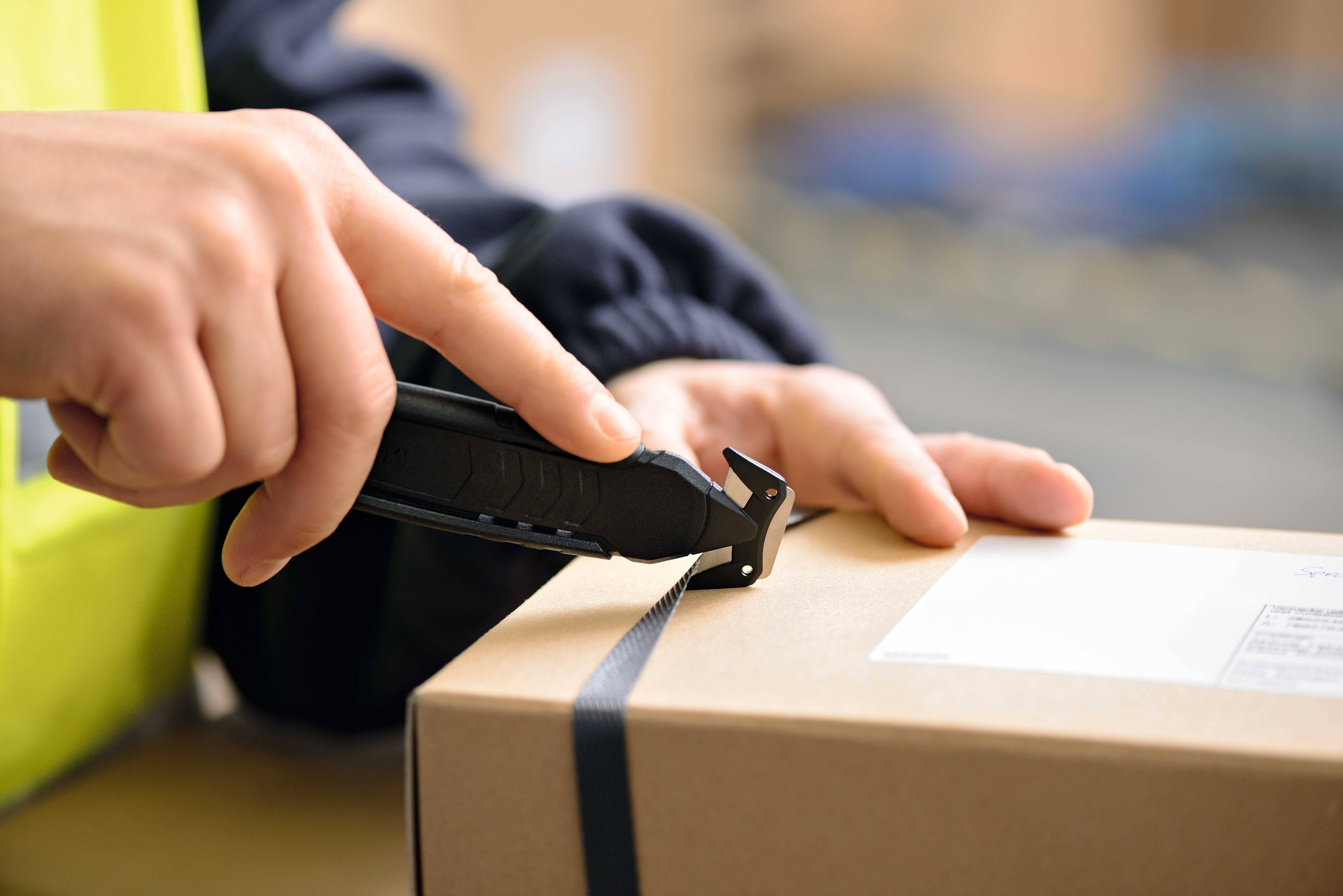 Osoba ostrożnie otwiera karton nożem do cięcia, który jest zabezpieczony czarną taśmą klejącą.