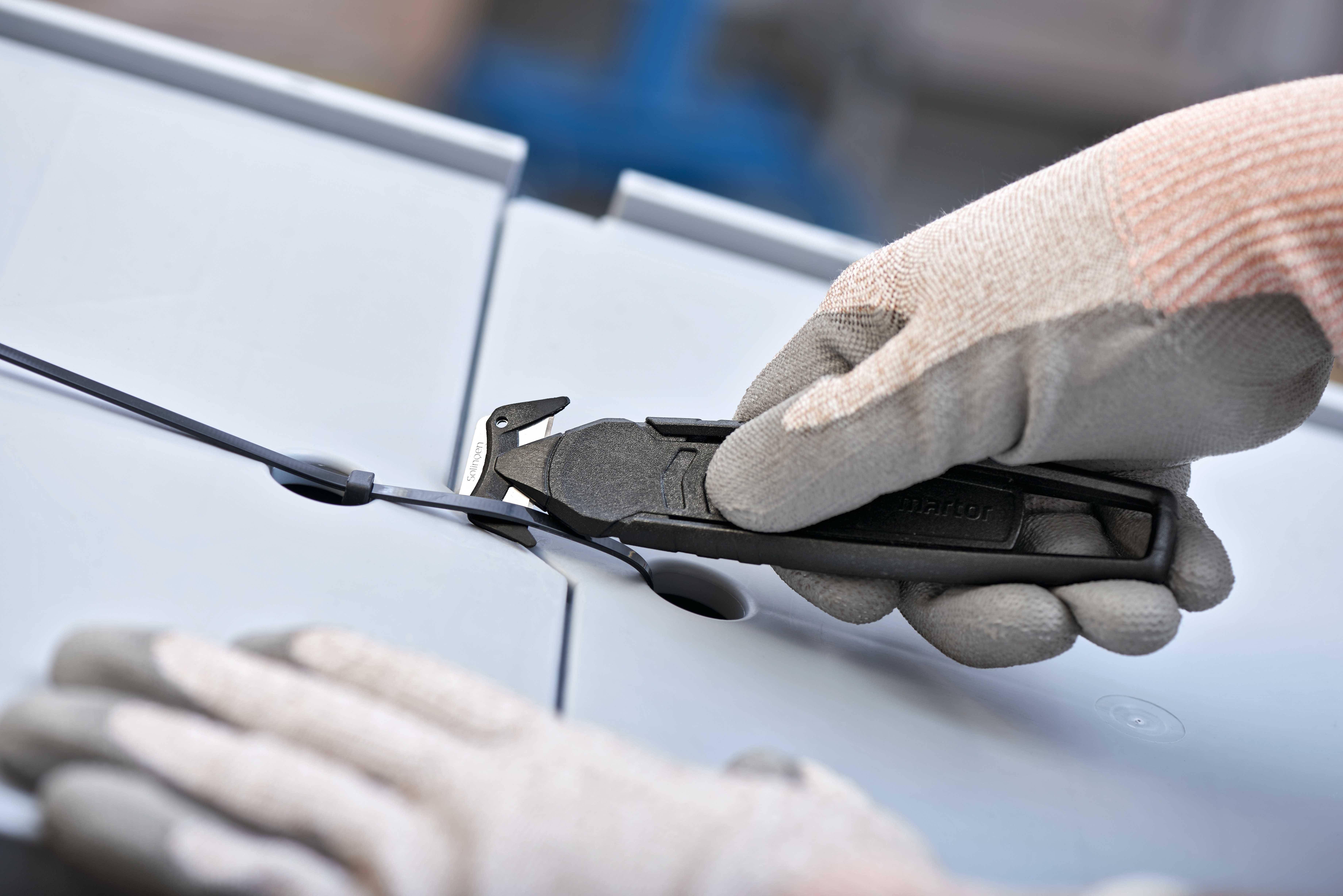 Dłoń w rękawicy trzyma obcinacz do drutu i przecina kabel na szarym tle.