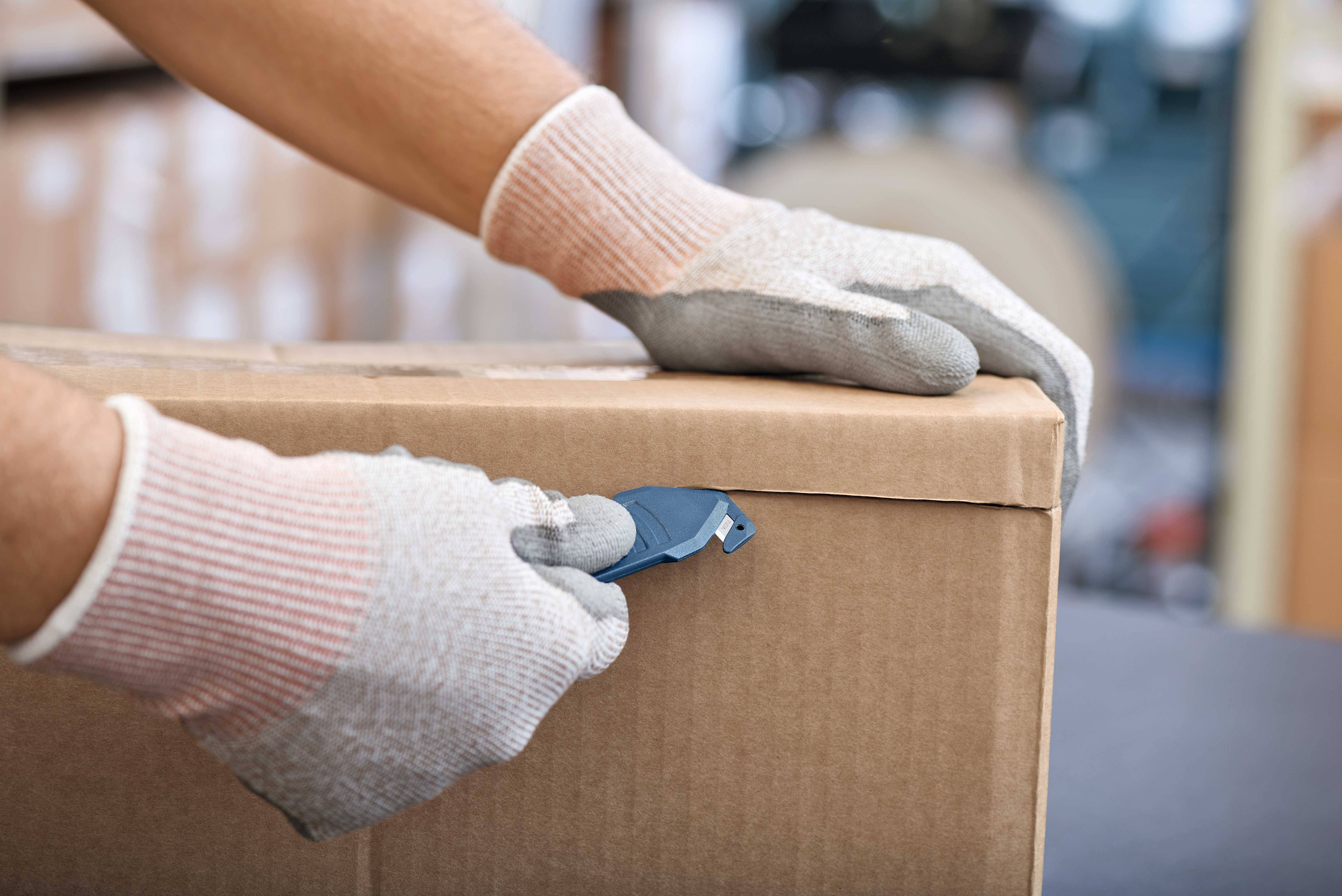 Osoba otwiera karton tekturowy za pomocą noża introligatorskiego. Ręce są zabezpieczone rękawicami roboczymi.