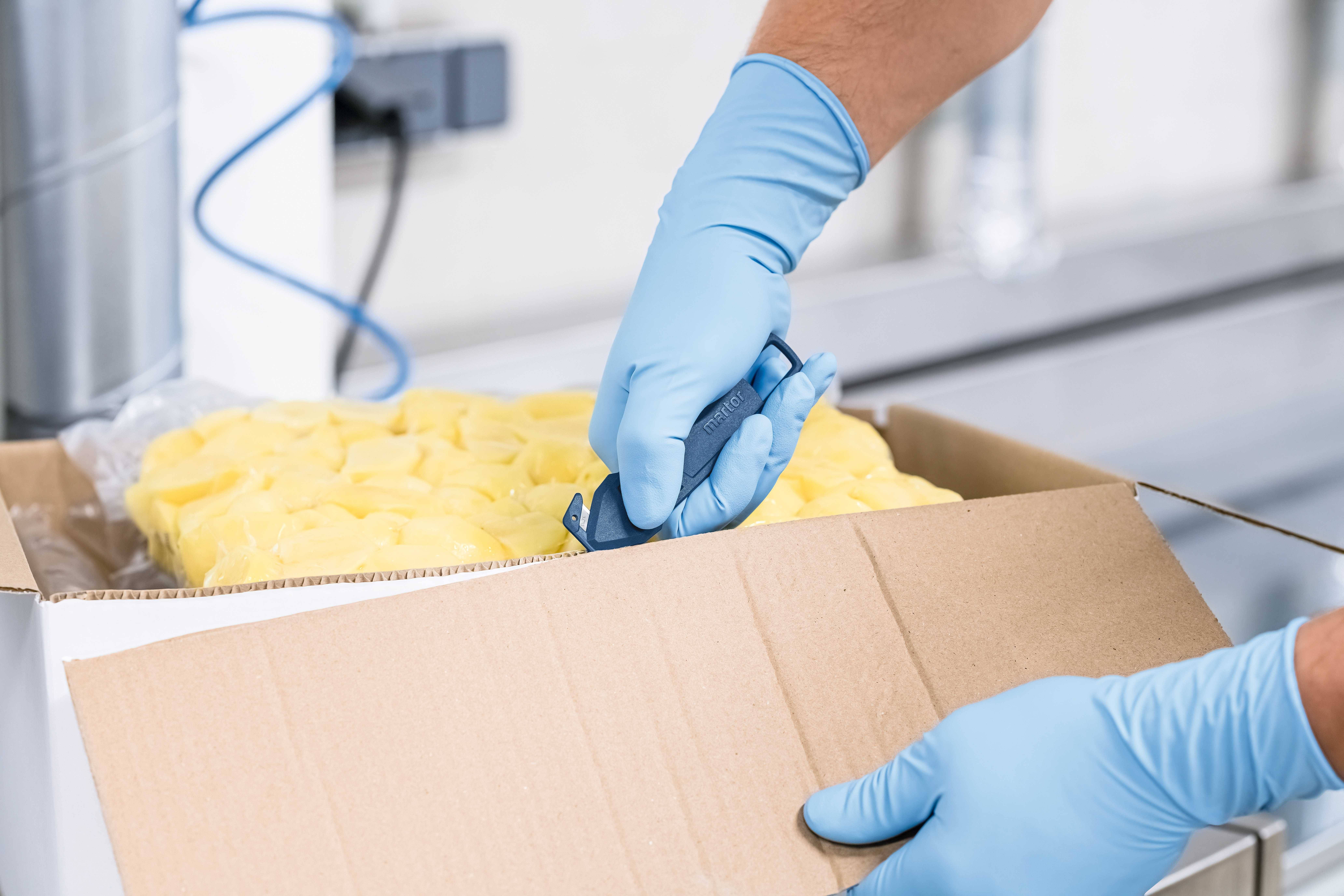 Osoba rozcina karton nożem. Osoba nosi niebieskie rękawice ochronne. Wewnątrz kartonu widoczne są żółte opakowania.