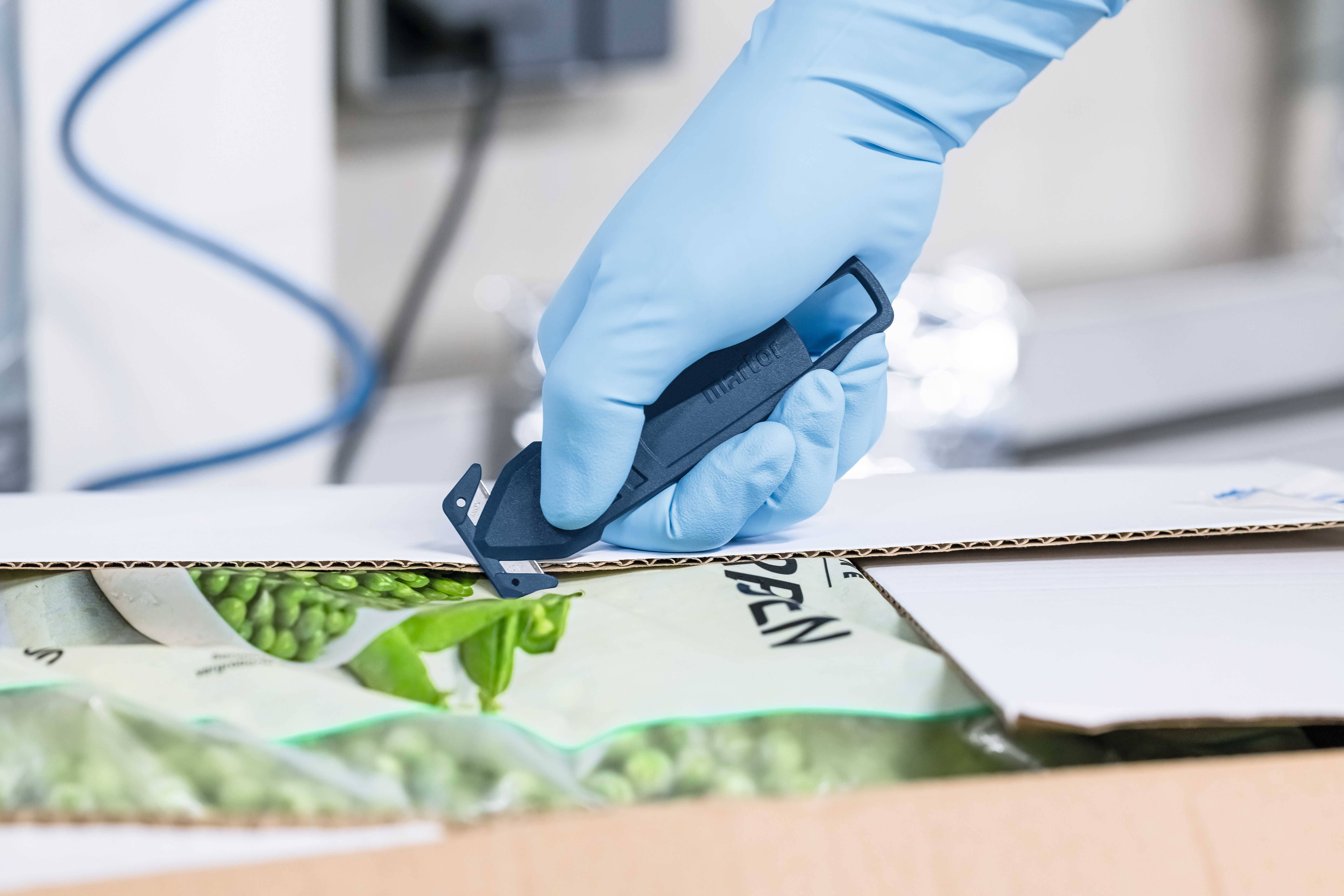 Dłoń w niebieskiej rękawiczce otwiera nożem karton z mrożonymi warzywami.