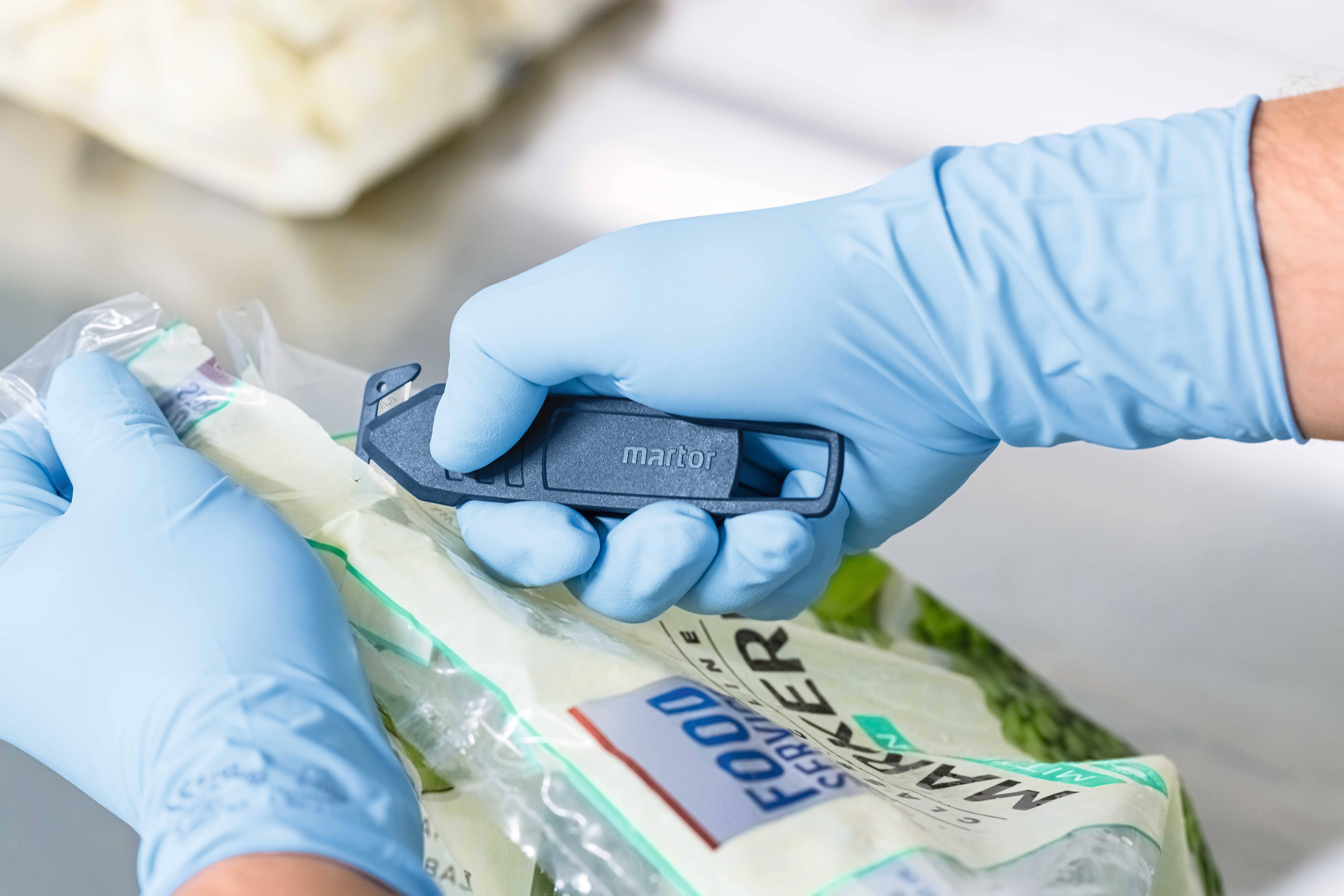 Osoba w niebieskich rękawiczkach otwiera opakowanie plastikowe niebieskim nożem bezpieczeństwa w czystym otoczeniu.