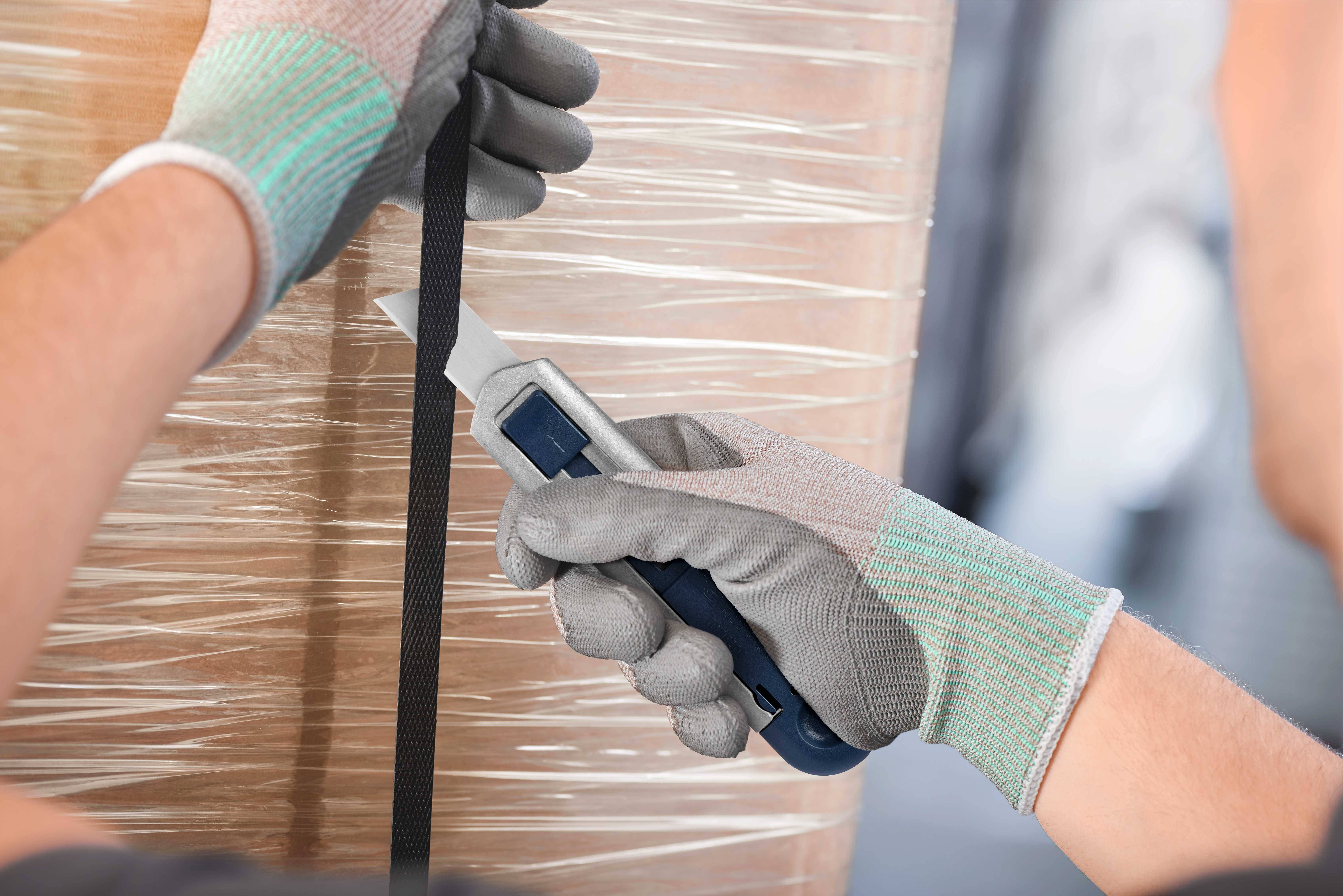 Zbliżenie rąk w rękawicach ochronnych używających noża bezpiecznego do przecięcia czarnej plastikowej taśmy na palecie.