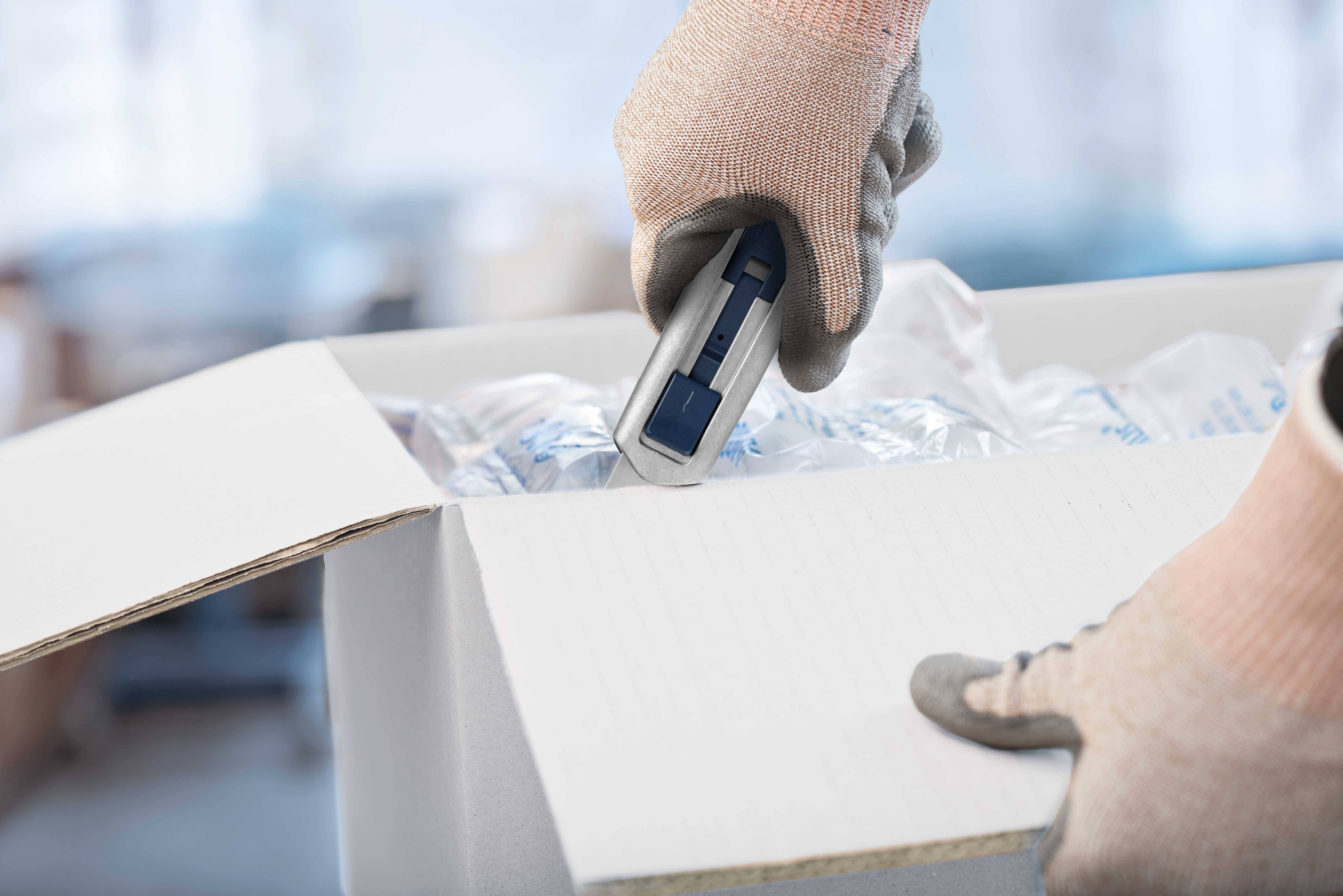 Osoba w rękawiczkach otwiera karton za pomocą noża do cięcia. Widoczny jest materiał ochronny zabezpieczający zawartość.