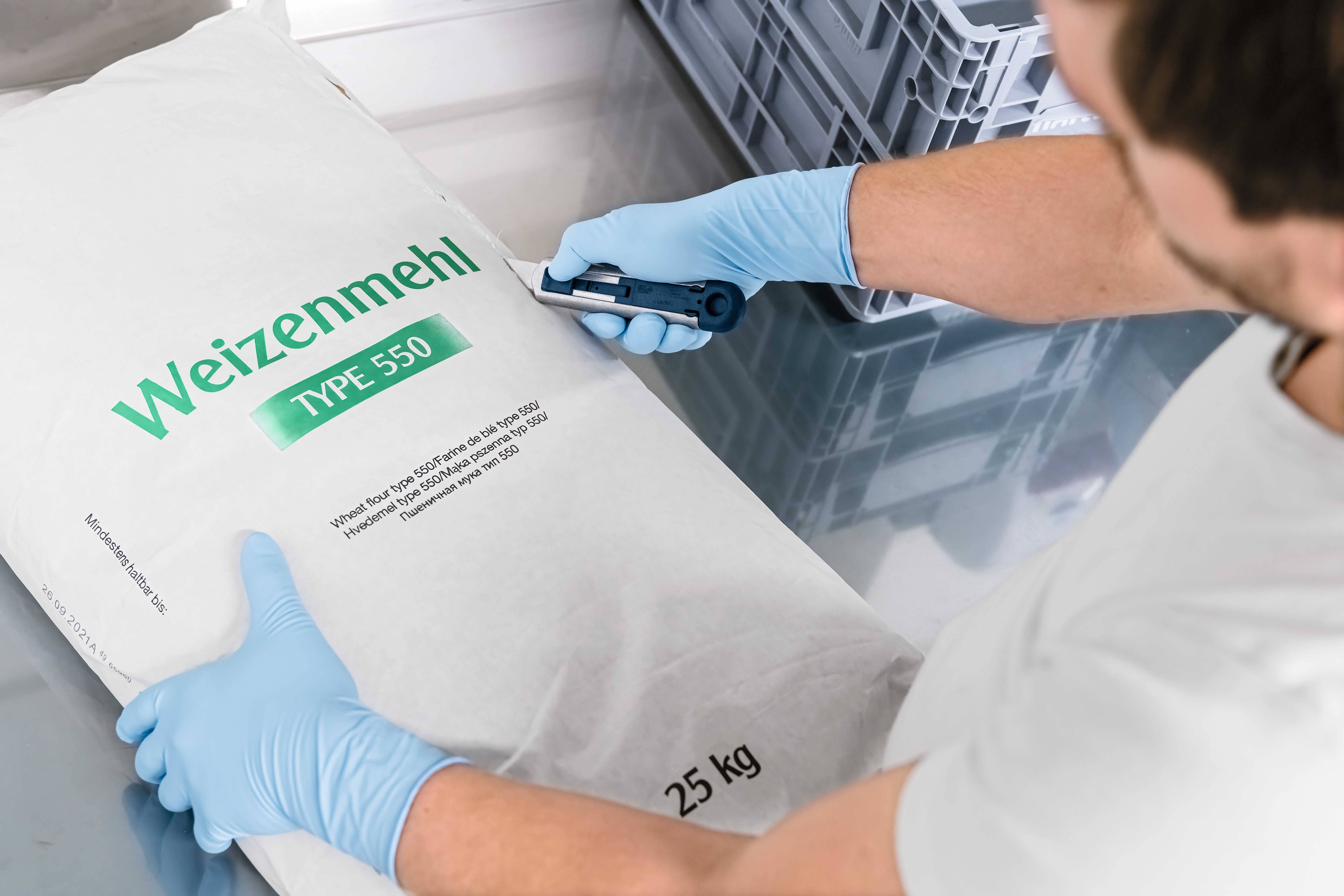 Mężczyzna w niebieskich rękawiczkach otwiera 25-kilogramowy worek mąki pszennej typ 550 za pomocą noża monterskiego.