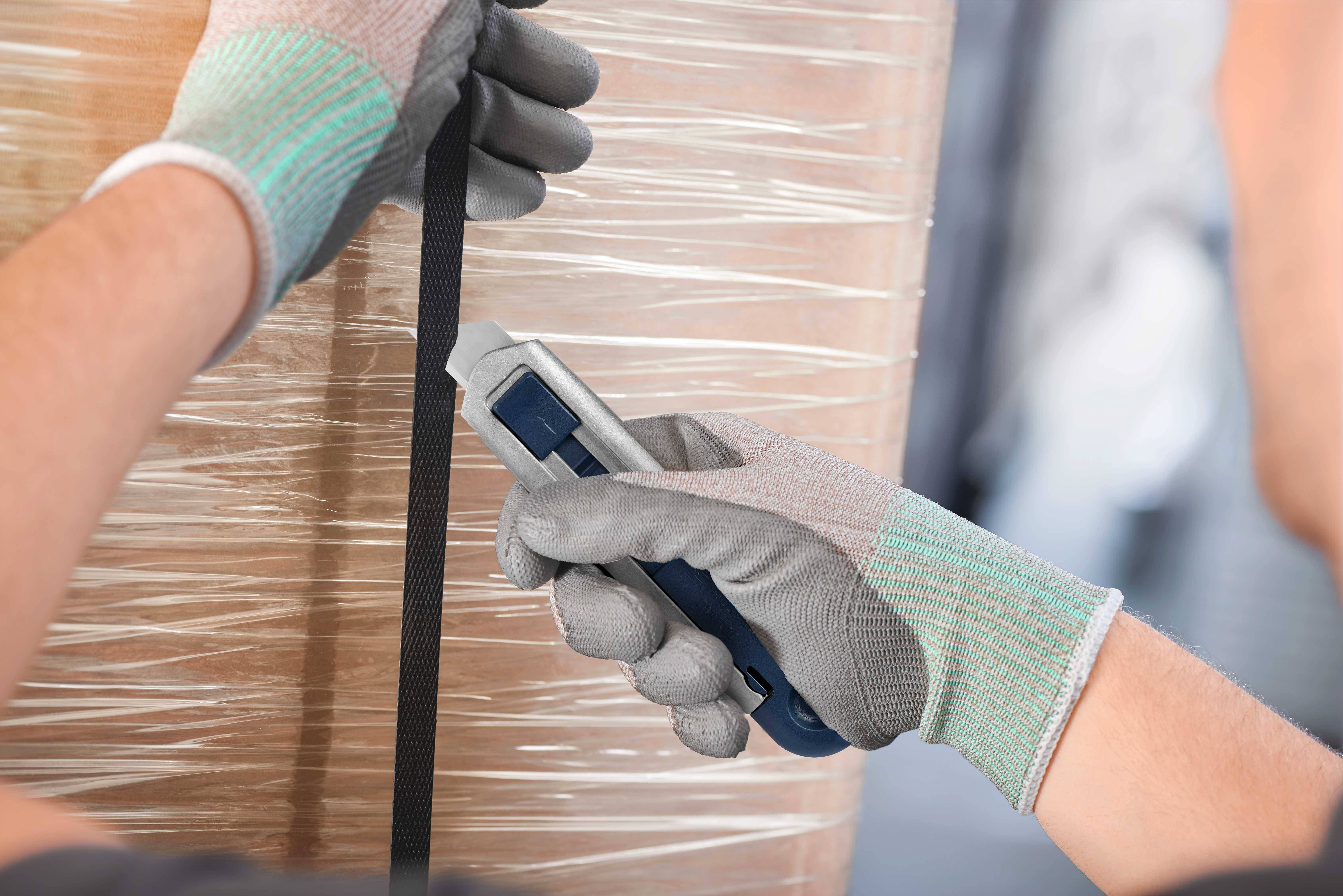 Osoba w rękawiczkach przecina nożem czarną taśmę na palecie owiniętej folią.