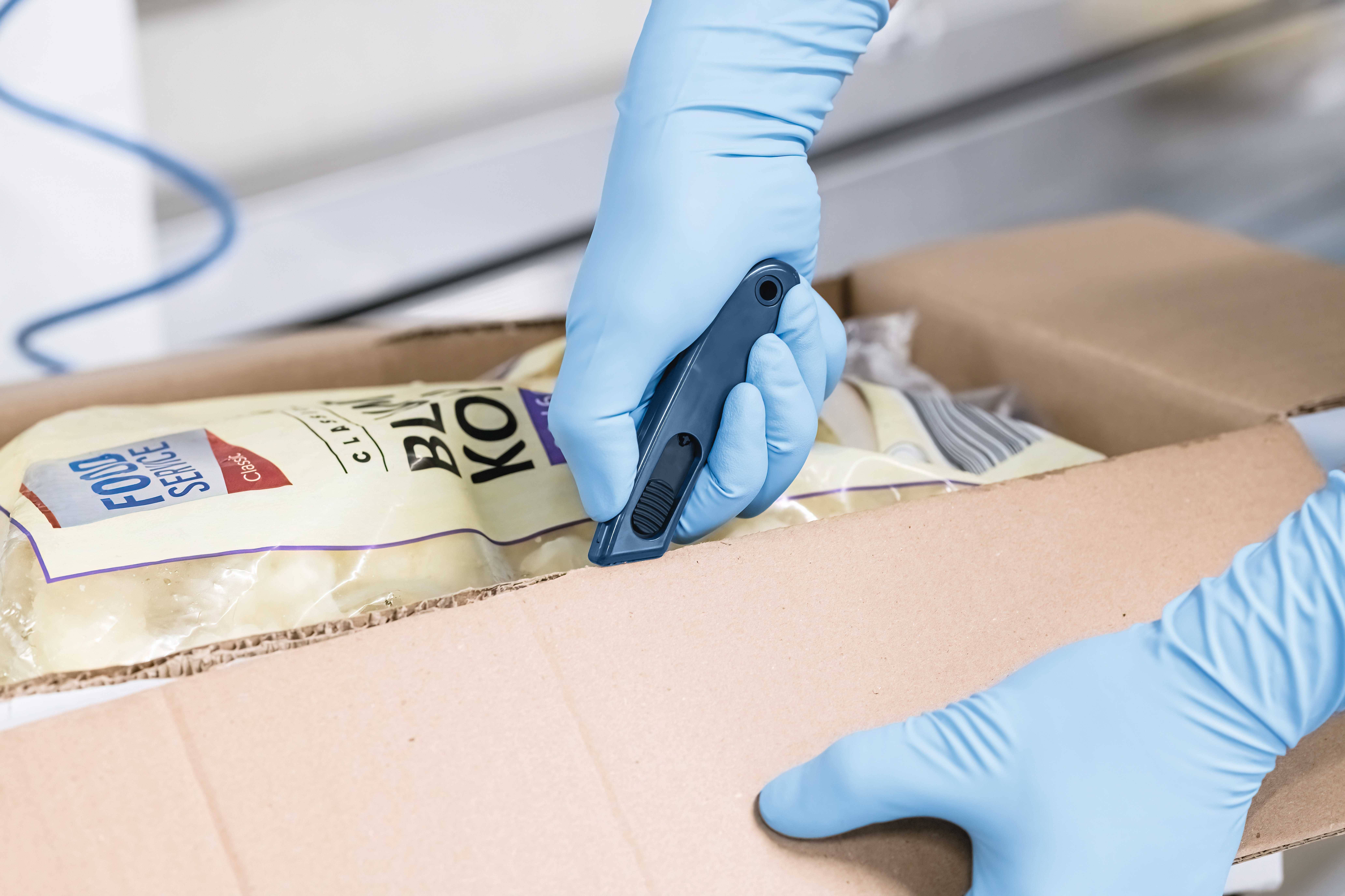Osoba ostrożnie otwiera karton za pomocą noża do cięcia. W kartonie znajdują się produkty pakowane próżniowo, co widać po etykietach.
