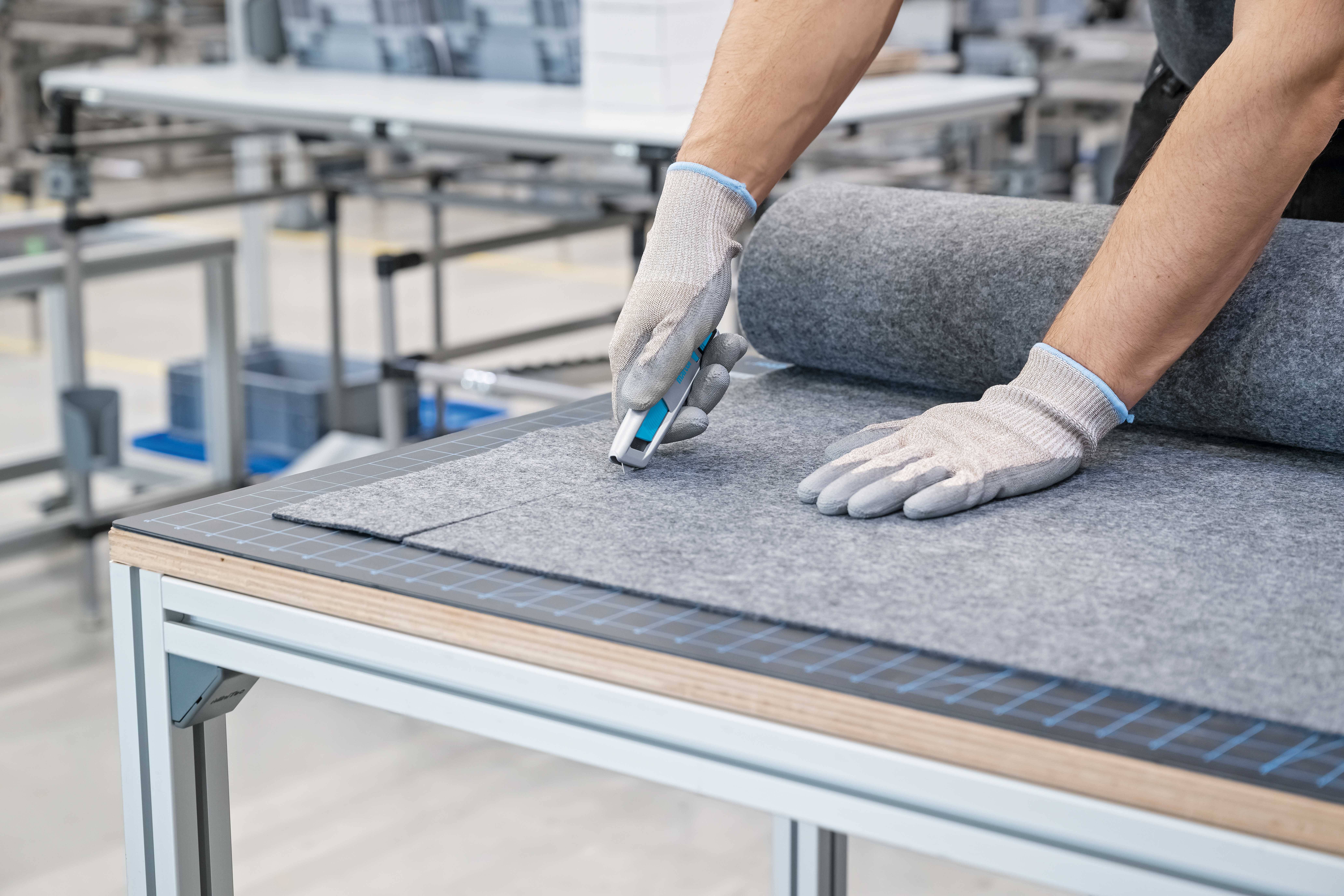 Robotnik w rękawicach tnie nożem introligatorskim szary materiał na stole w fabryce.