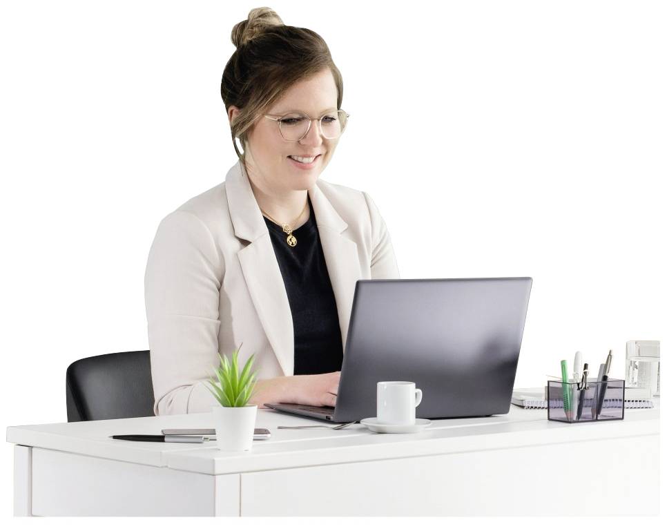 Osoba siedzi przy biurku, pracuje na laptopie i się uśmiecha. Na stole stoją kubek kawy, roślina oraz przybory do pisania.