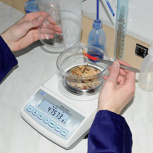 Dwie ręce układają kawałki drewna w szklanym pojemniku na wadze elektronicznej. Wyświetlacz wskazuje 45,73 g.