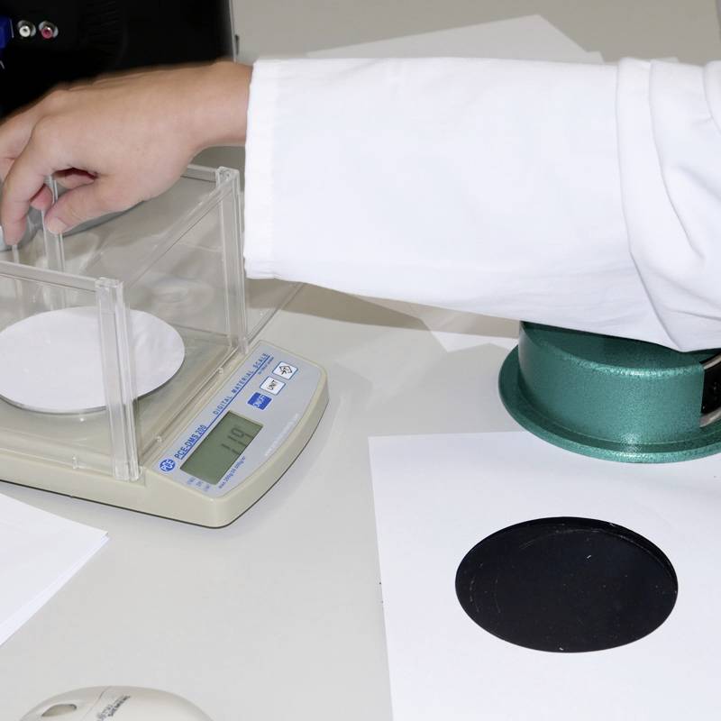 Dłoń w białym kitlu laboratoryjnym obsługuje cyfrową wagę z małą białą płytką, obok której znajduje się zielony obiekt.