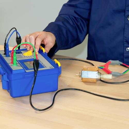 Osoba testuje urządzenie elektroniczne za pomocą różnych kabli i przyrządu pomiarowego na stole.