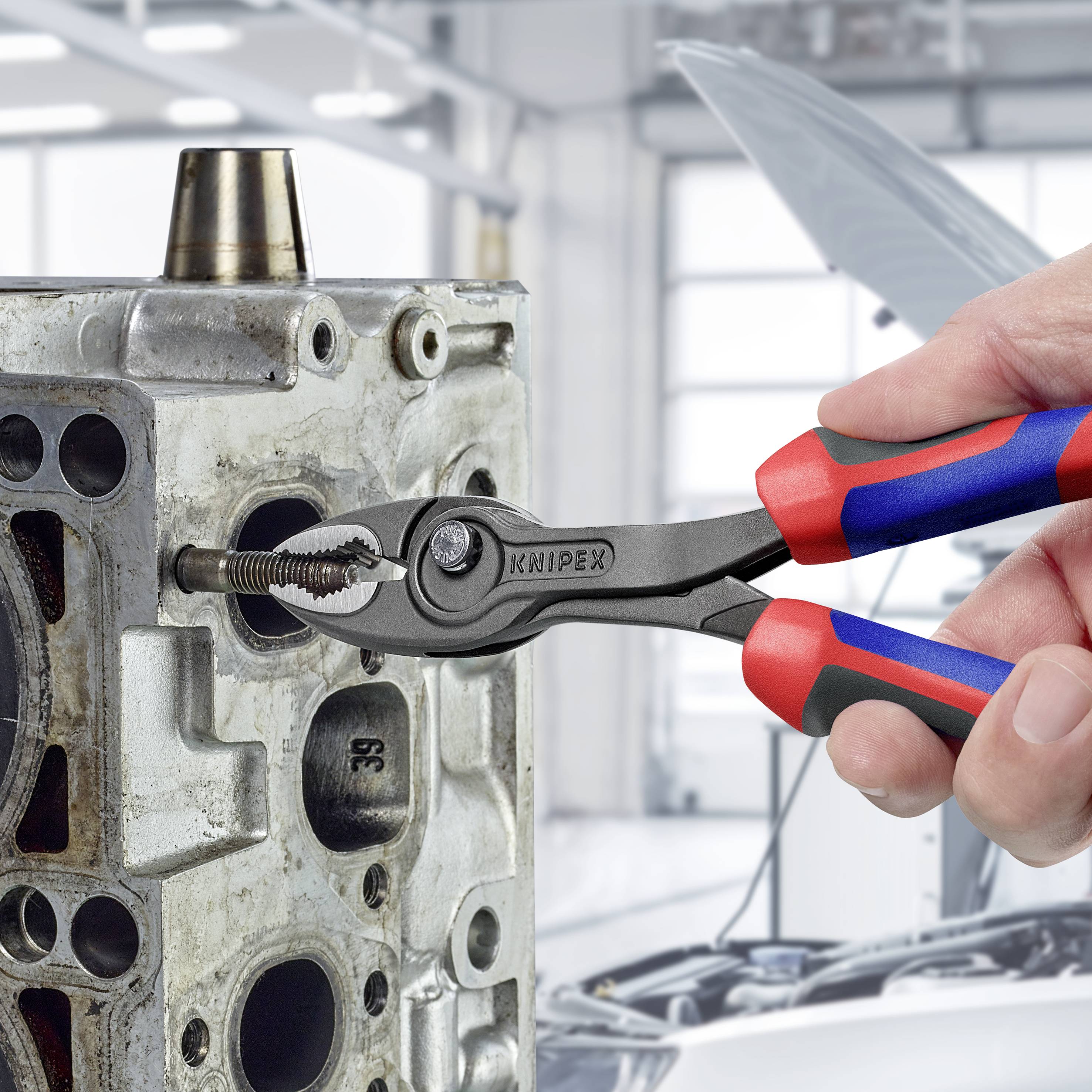 Osoba trzyma szczypce podczas pracy przy bloku silnika. W tle widać wnętrze warsztatu samochodowego.