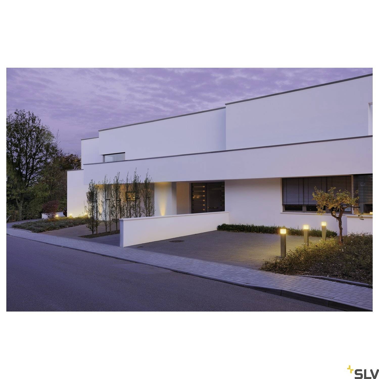 Nowoczesny biały budynek z płaskim dachem, otoczony wybrukowanymi ścieżkami i drzewami. Oświetlone lampy zewnętrzne wzdłuż chodnika.