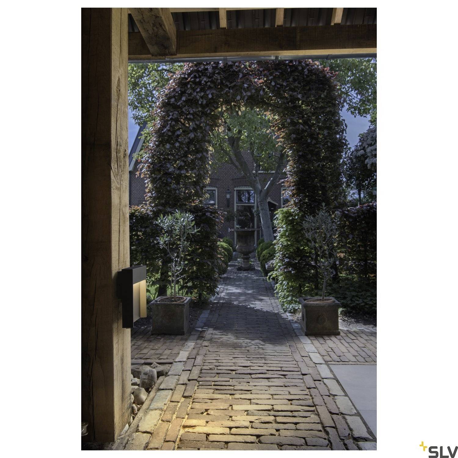 Brukowana ścieżka prowadzi przez łuk utworzony z gęstych liści, otoczony bujną zielenią. W tle widać budynek z czerwonej cegły.