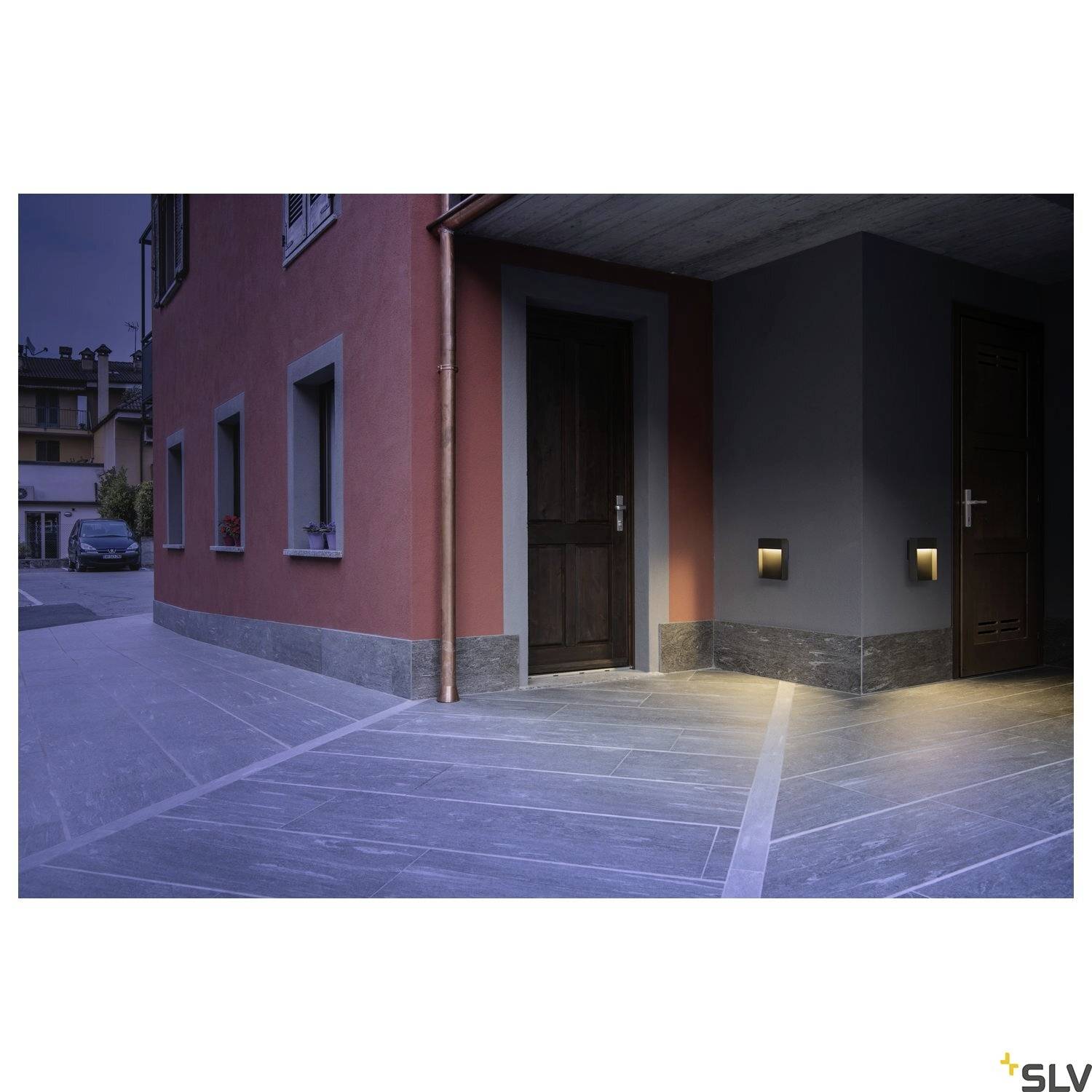 Widok z zewnątrz domu o zmierzchu z oświetlonym wejściem. Lampy na ścianie zewnętrznej oświetlają chodnik.