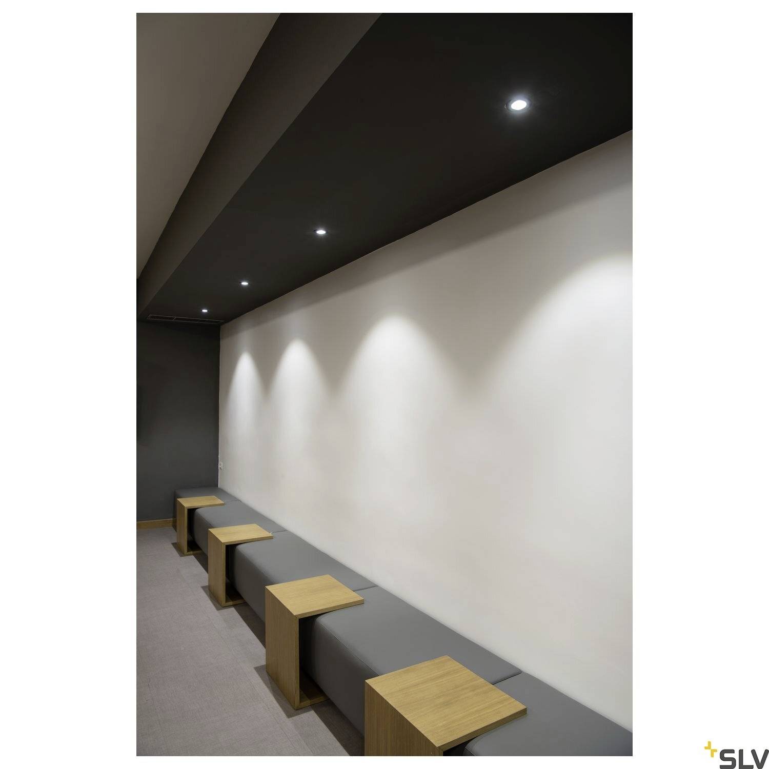 Nowoczesne pomieszczenie z szaro-białymi ścianami, zintegrowanymi sufitowymi reflektorami oraz minimalistycznymi drewnianymi stołkami ustawionymi wzdłuż ściany.