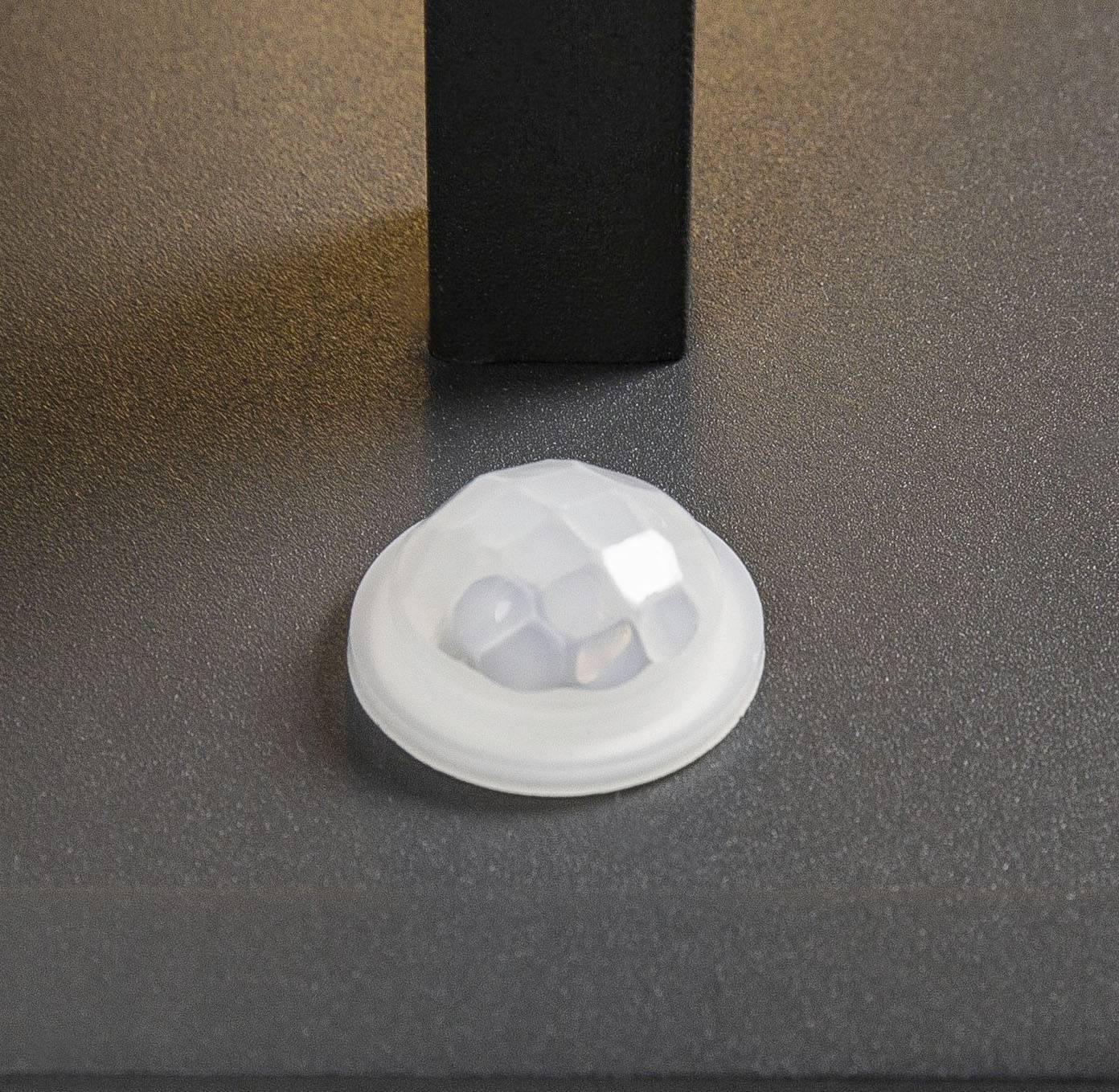 Czarna noga stołu stoi na gładkiej, szarej podłodze. Obok leży biała, sześciokątna gumowa stopka, która nie jest zamontowana.