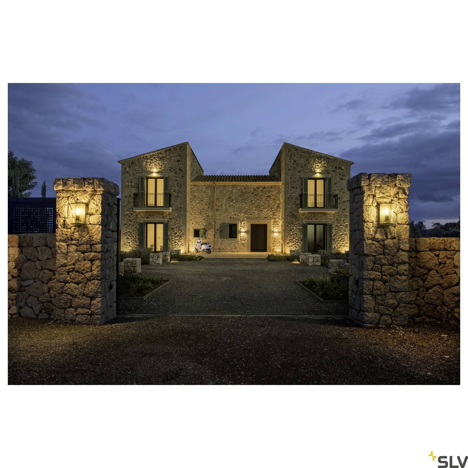 Oświetlony kamienny dom o zmierzchu, otoczony kamiennymi murami, z dwoma latarniami przy wejściu. Światła domów są włączone.