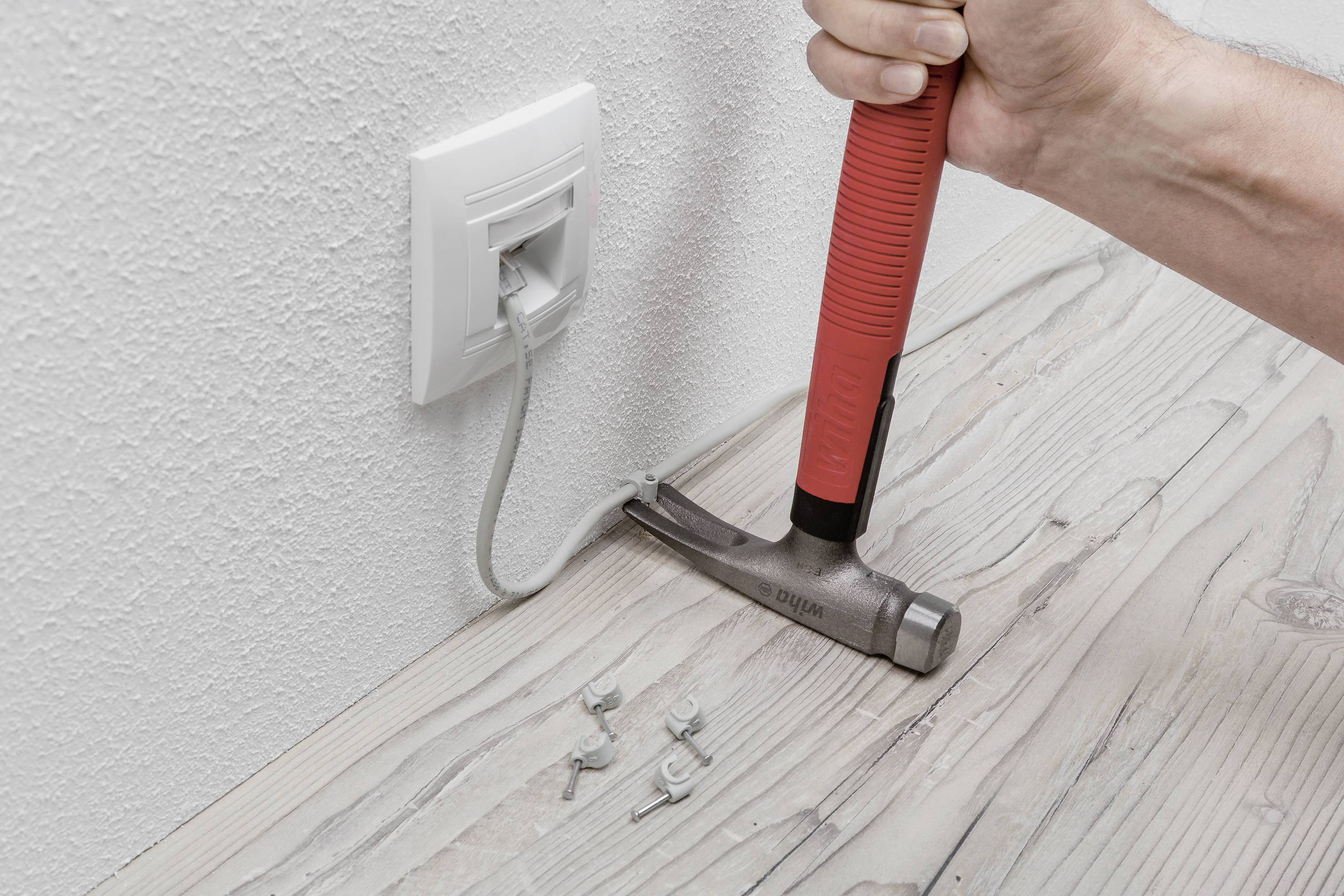 Ręka z młotkiem usuwa klamry mocujące kabel z drewnianej podłogi. Kabel sieciowy jest podłączony do gniazdka ściennego.