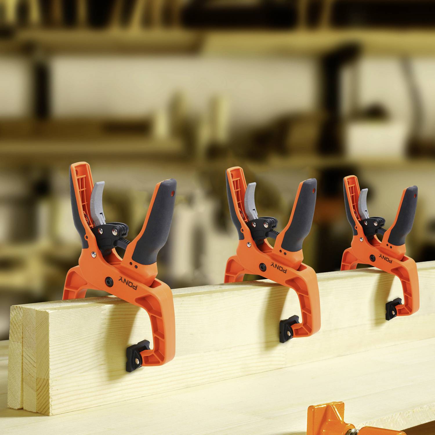 Trzy pomarańczowe zaciski przytrzymują deskę drewnianą w warsztacie. W tle widać rozmyte kolejne zaciski.