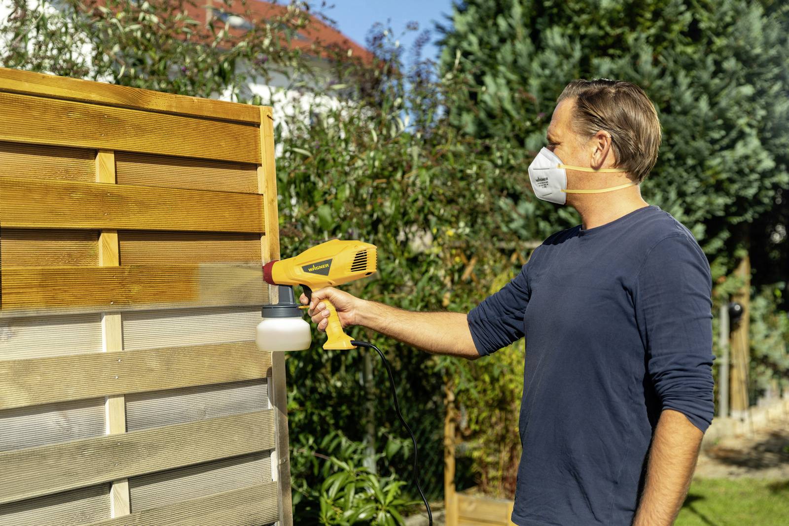 Mężczyzna w masce nakłada środek ochronny na drewniany płot na wolnym powietrzu. W tle widać drzewa i dach.