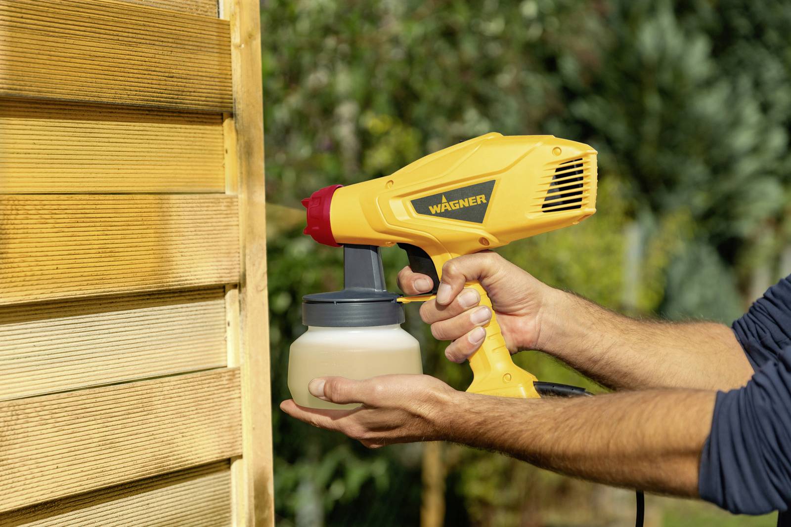 Człowiek używa żółtego urządzenia natryskowego do lakierowania drewnianej ściany. Słońce świeci, a tło jest rozmyte, w tle widać drzewa.