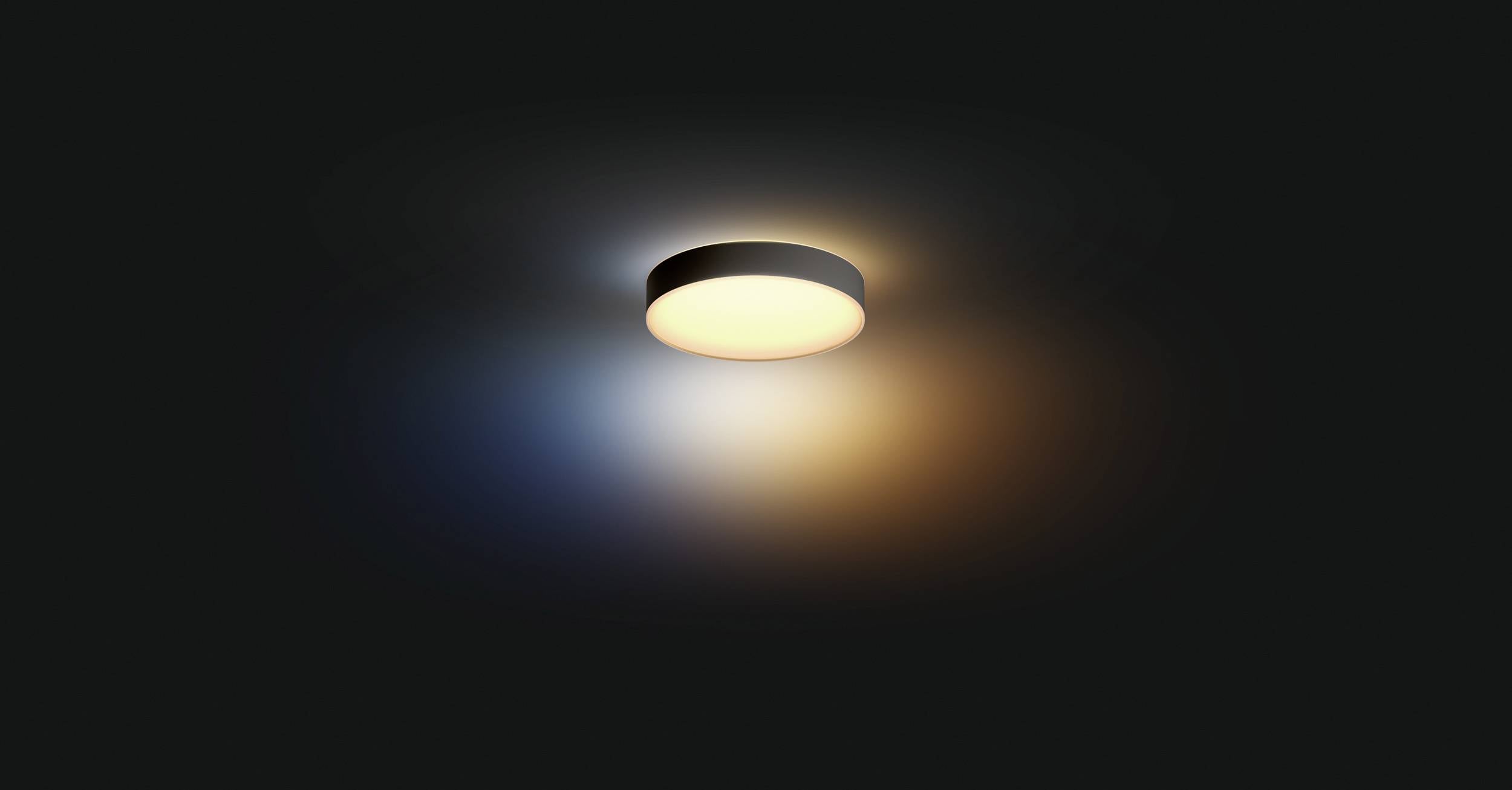 Okrągła lampa sufitowa rzuca ciepłe, żółtawe światło w ciemnym pomieszczeniu. Delikatny gradient barw niebieskiego i pomarańczowego otacza ją wokoło.