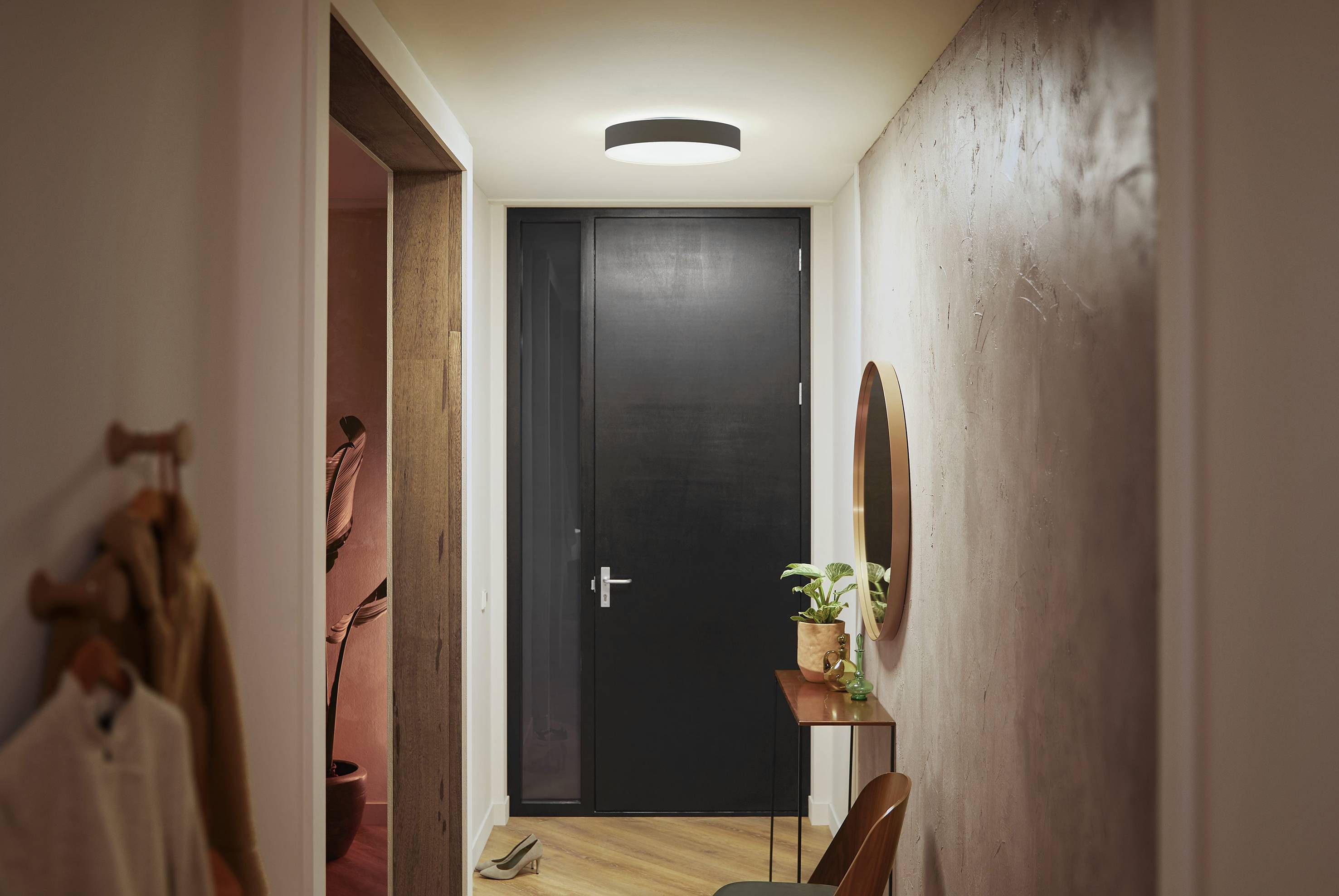 Korytarz z jasną lampą sufitową, czarnymi drzwiami na końcu, małym stolikiem z rośliną oraz okrągłym lustrem na ścianie.