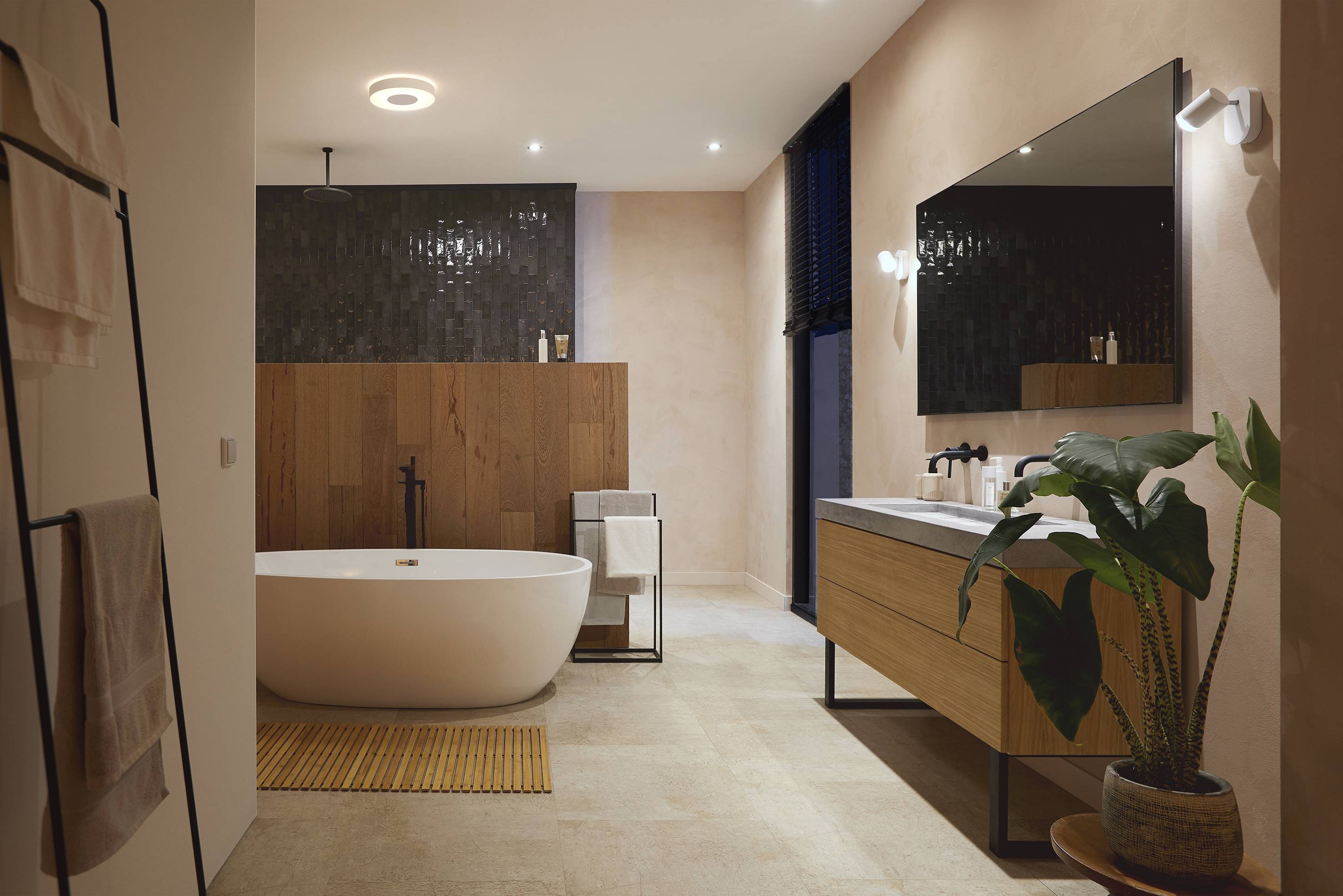 Nowoczesna łazienka ze swobodnie stojącą wanną, drewnianą zabudową, dużym lustrem i umywalką; elementy roślinne i dekoracyjne dopełniają projekt.