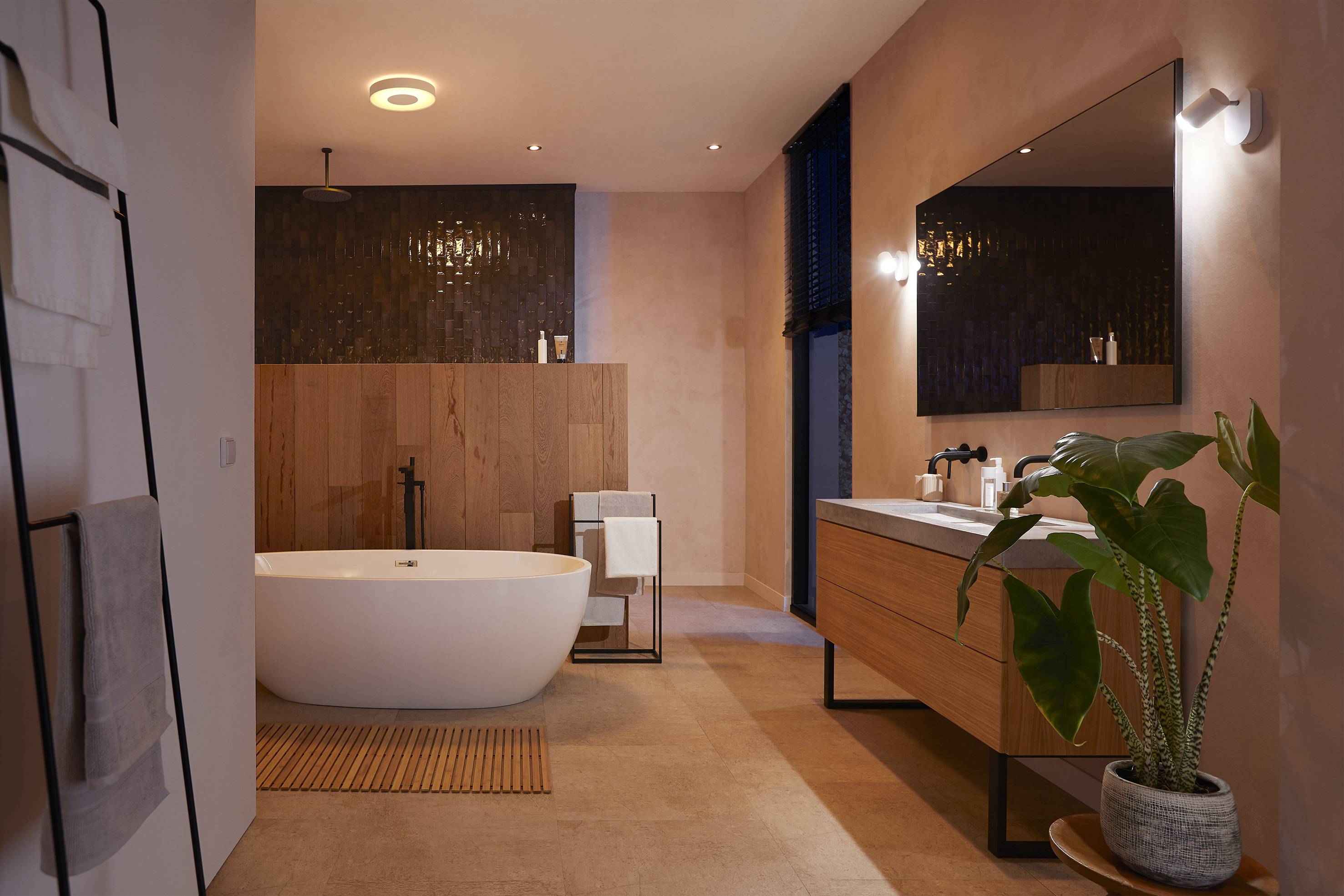 Nowoczesna łazienka z wolnostojącą wanną, drewnianą boazerią, dużym lustrem i rośliną na podłodze, w nastrojowym oświetleniu.