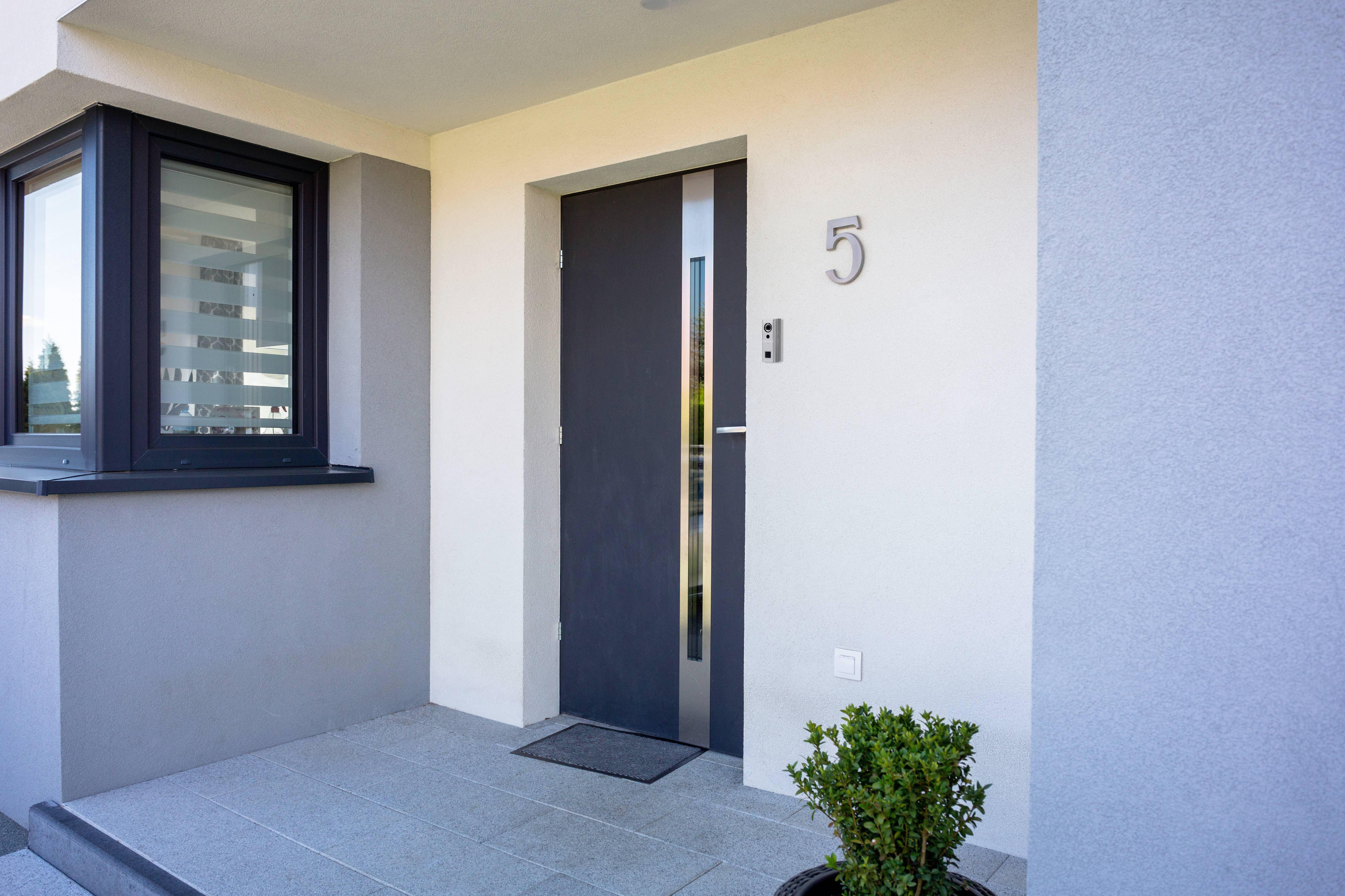 Wejście do nowoczesnego domu z szarymi drzwiami, numerem domu 5 i małą doniczką z rośliną po prawej stronie drzwi.